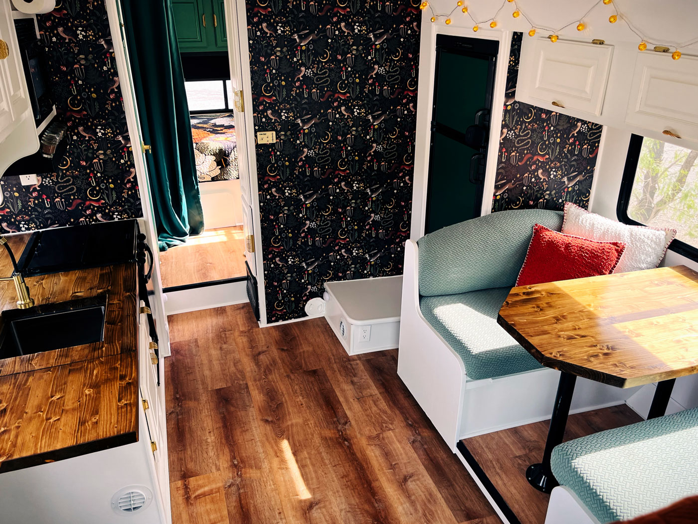 Cozy RV interior featuring a wooden kitchen counter, patterned wallpaper, cushioned bench seating with red cushions, and a small dining table beside a sunlit window.