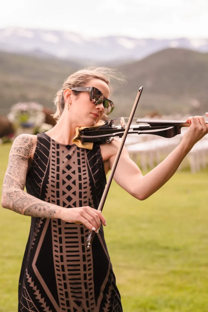 A musician with tattooed arms and sunglasses plays an electric violin outdoors against a backdrop of mountains at a Chaparral Ranch Wedding.