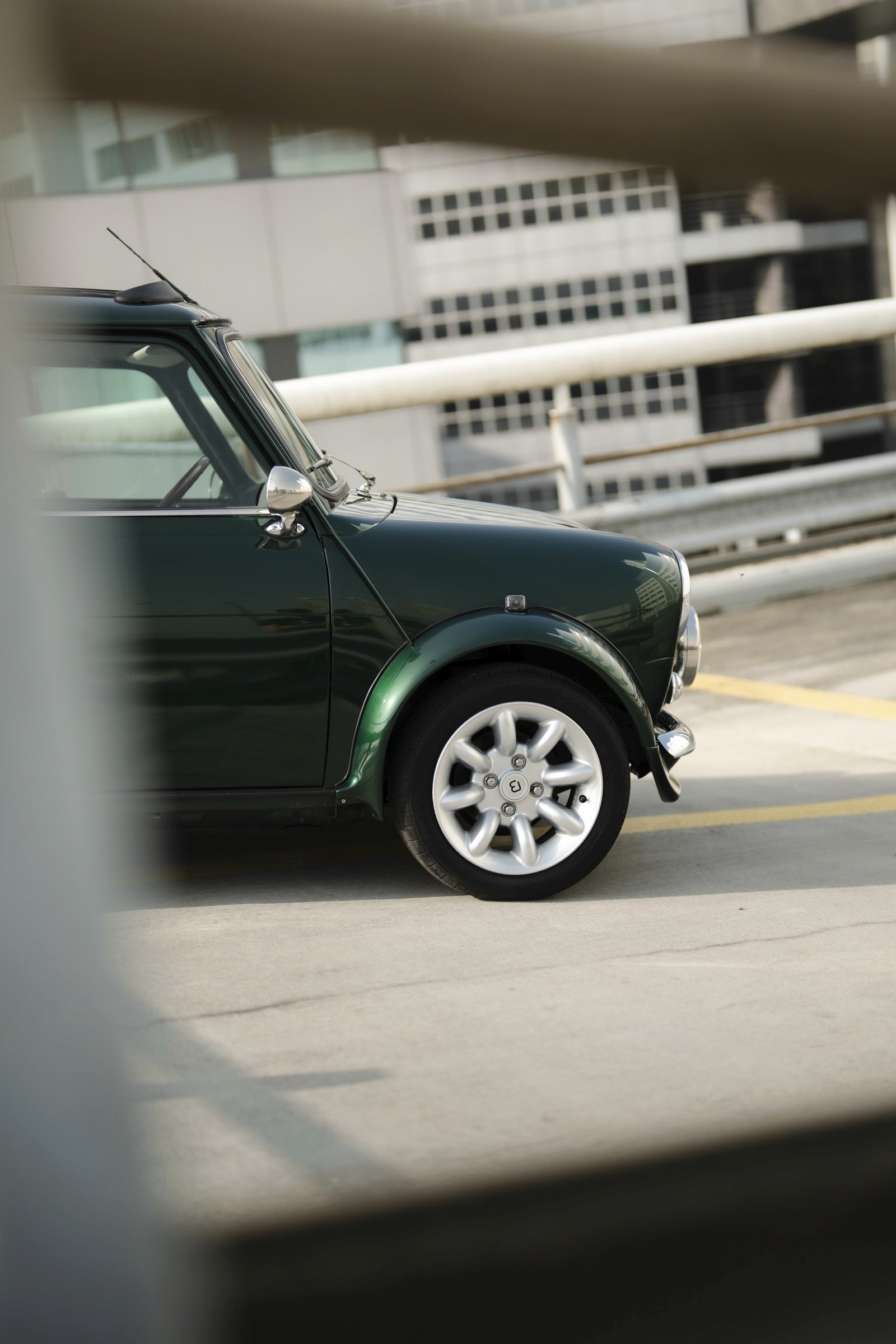 Close-up of a green car's wheel and side, partially obscured by a railing in the background.