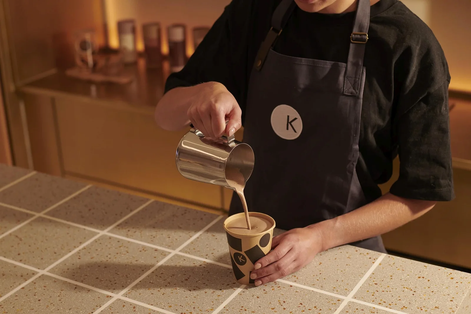 Barista pouring milk into branded coffee cup marked with "k" logo at counter.
