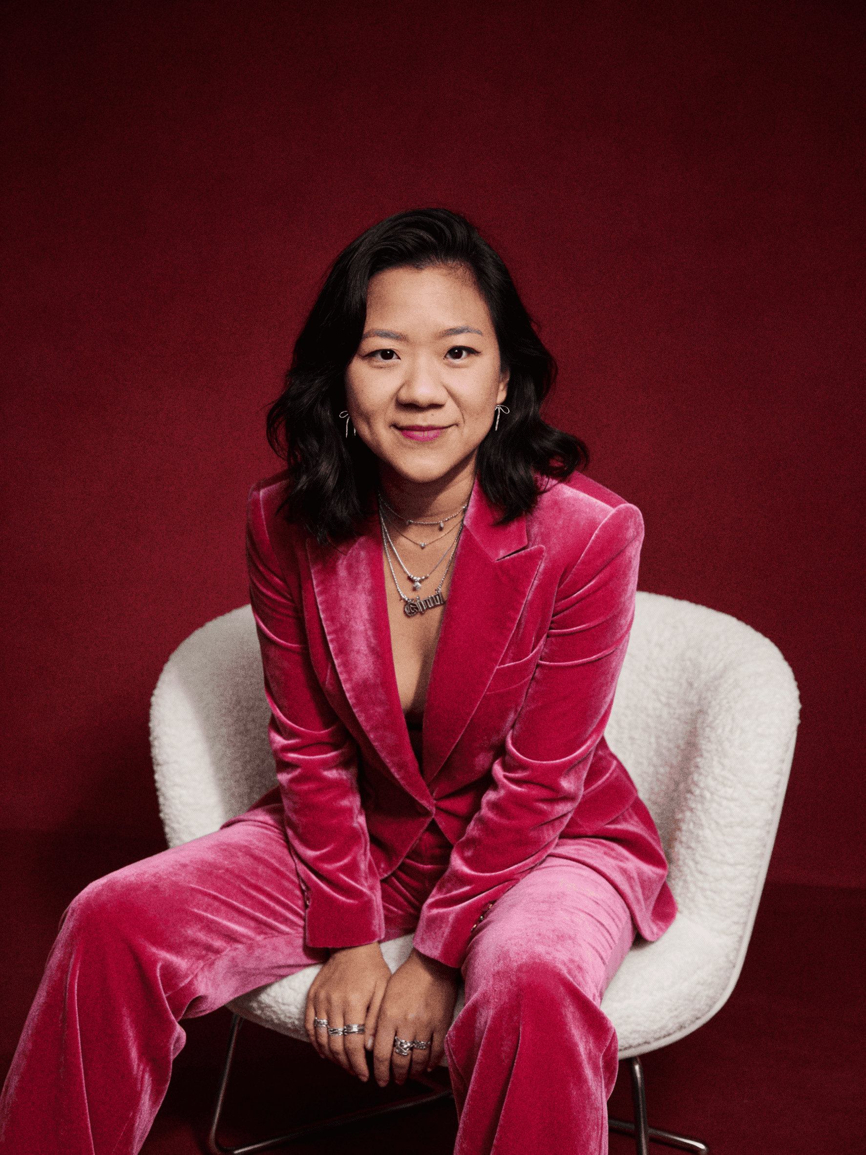 Studio portrait of Mikaela, a Vietnamese woman with dark, wavy medium length hair, wearing a pink velvet suit, sitting on a chair in front of a deep red background. She smiles confidently.