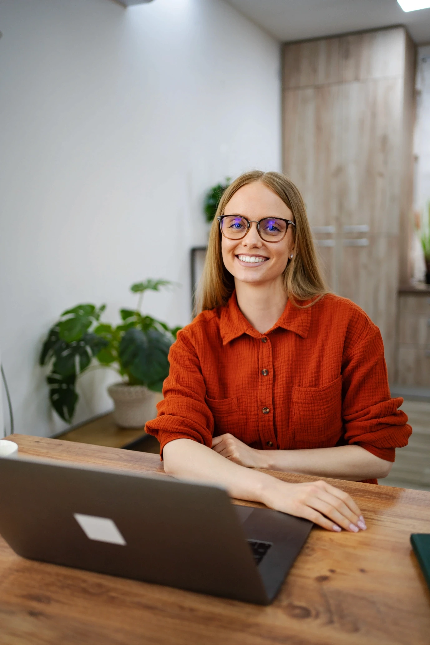 A woman with glasses and long hair sits at a wooden desk with a laptop in a modern, bright office space featuring plants and wooden cabinets.