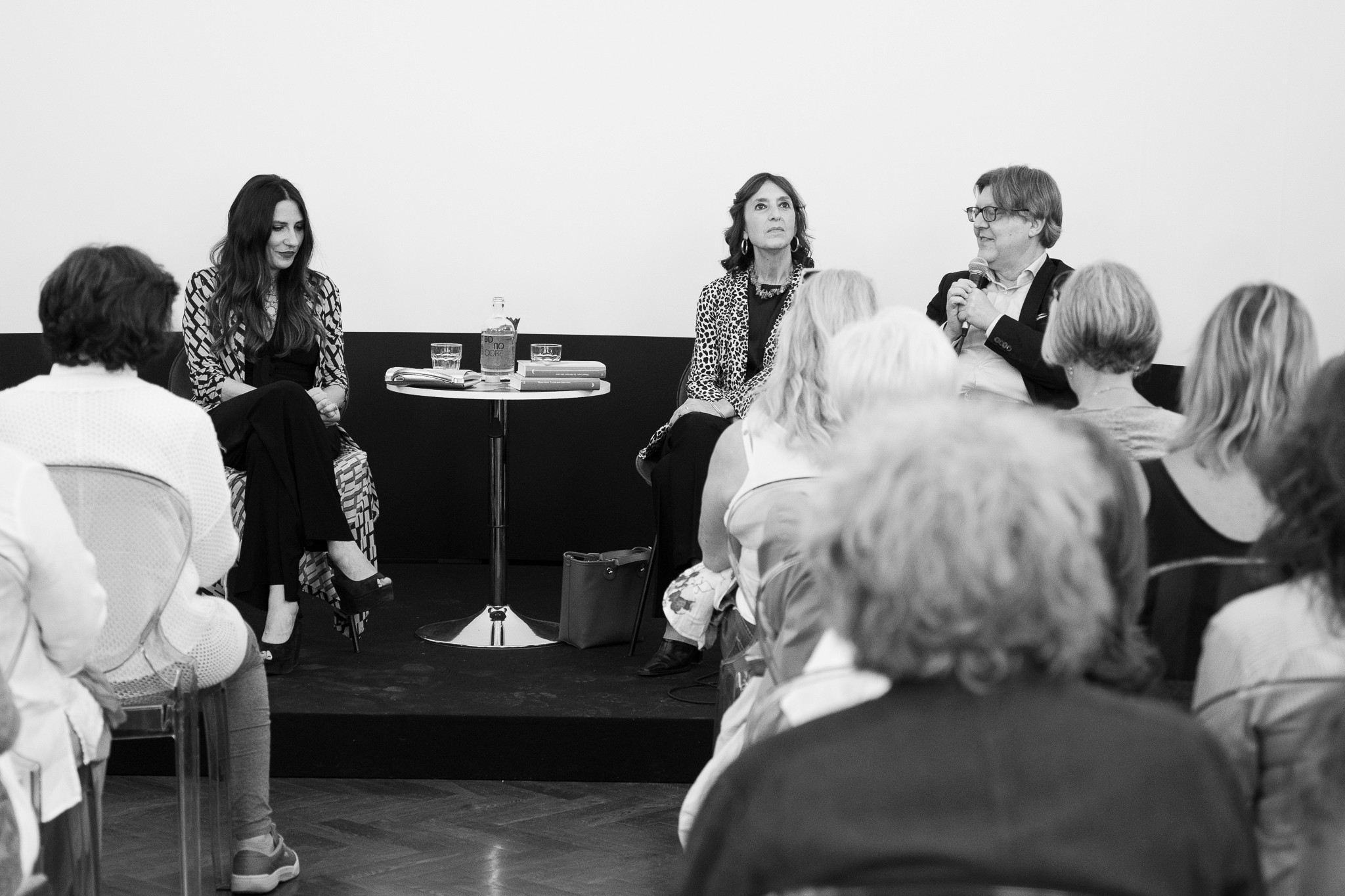 A young person with curly hair smiles at the camera while sitting in front of a laptop against a dark background.