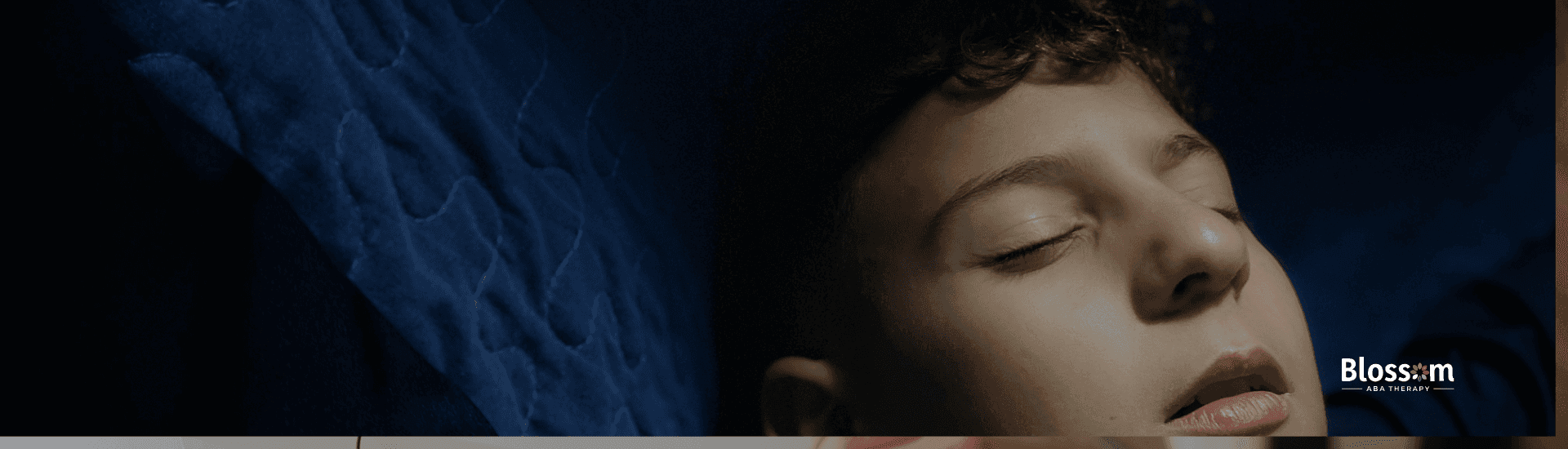 Boy lying under a dark blue blanket with his eyes closed and arms raised, appearing calm and relaxed in dim lighting.