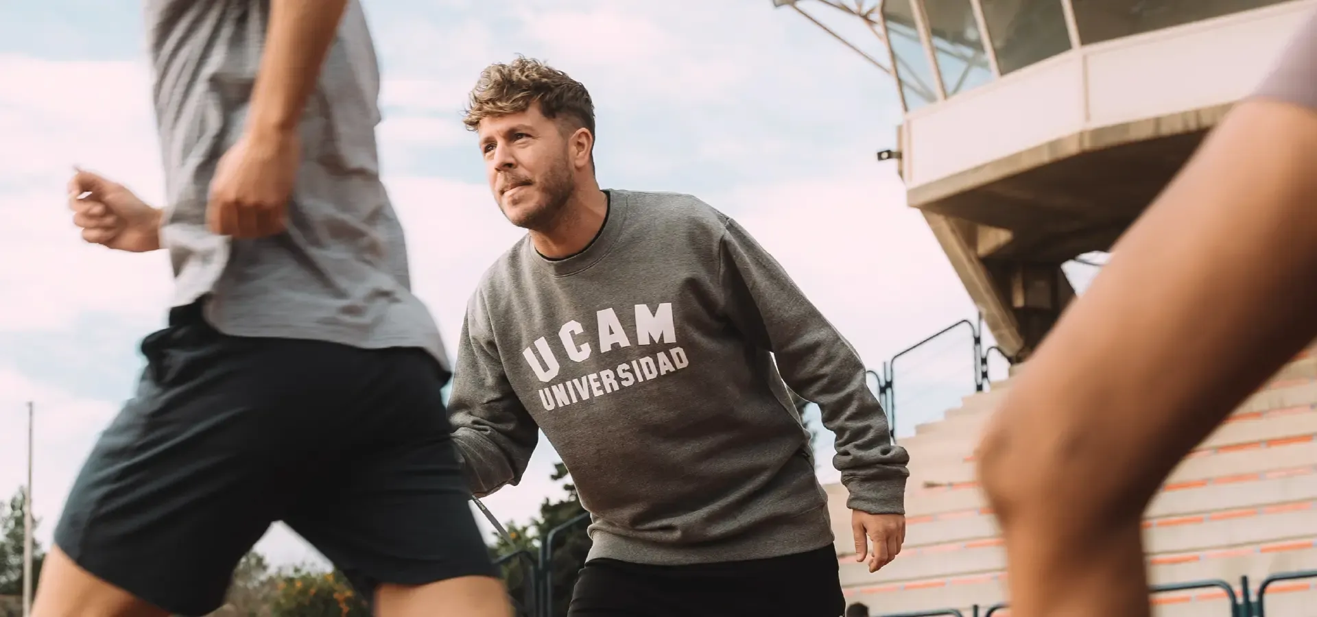 A person in a UCAM Universidad sweatshirt is actively engaged in a sports training session outdoors, with multiple individuals in athletic attire, emphasizing teamwork and fitness, relevant to European Master Sports Management.