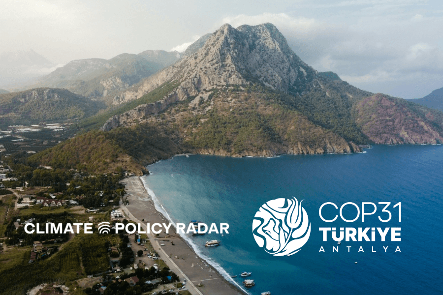 Aerial view of the Mediterranean coast near Antalya, Türkiye, where a steep, rocky mountain covered in pine forest drops down to a curved pebble beach and deep blue sea, with a small village and farmland along the shore. Overlaid in white are the Climate Policy Radar logo and the COP31 Türkiye Antalya logo.