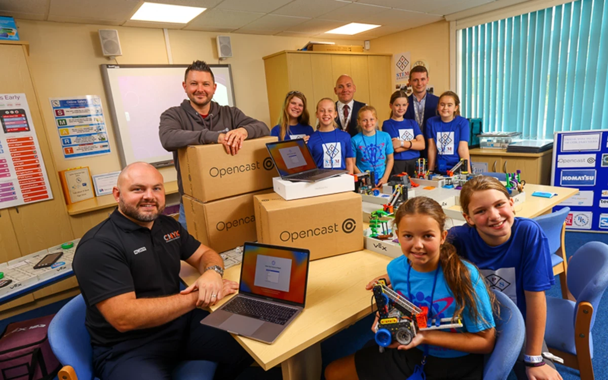A group of adults and children gather in a classroom around tables filled with robotics kits, laptops, and Opencast‑branded boxes, posing together during a STEM activity.