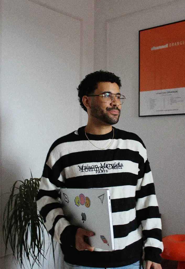 Karim standing near a plant in a minimalist studio space, smiling at the camera.