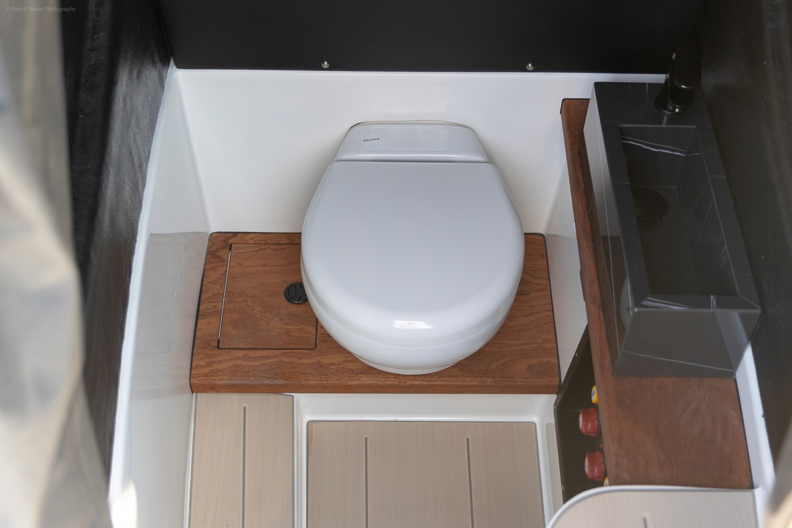 Compact yacht bathroom with white toilet on teak wood platform and built-in storage cabinets below.