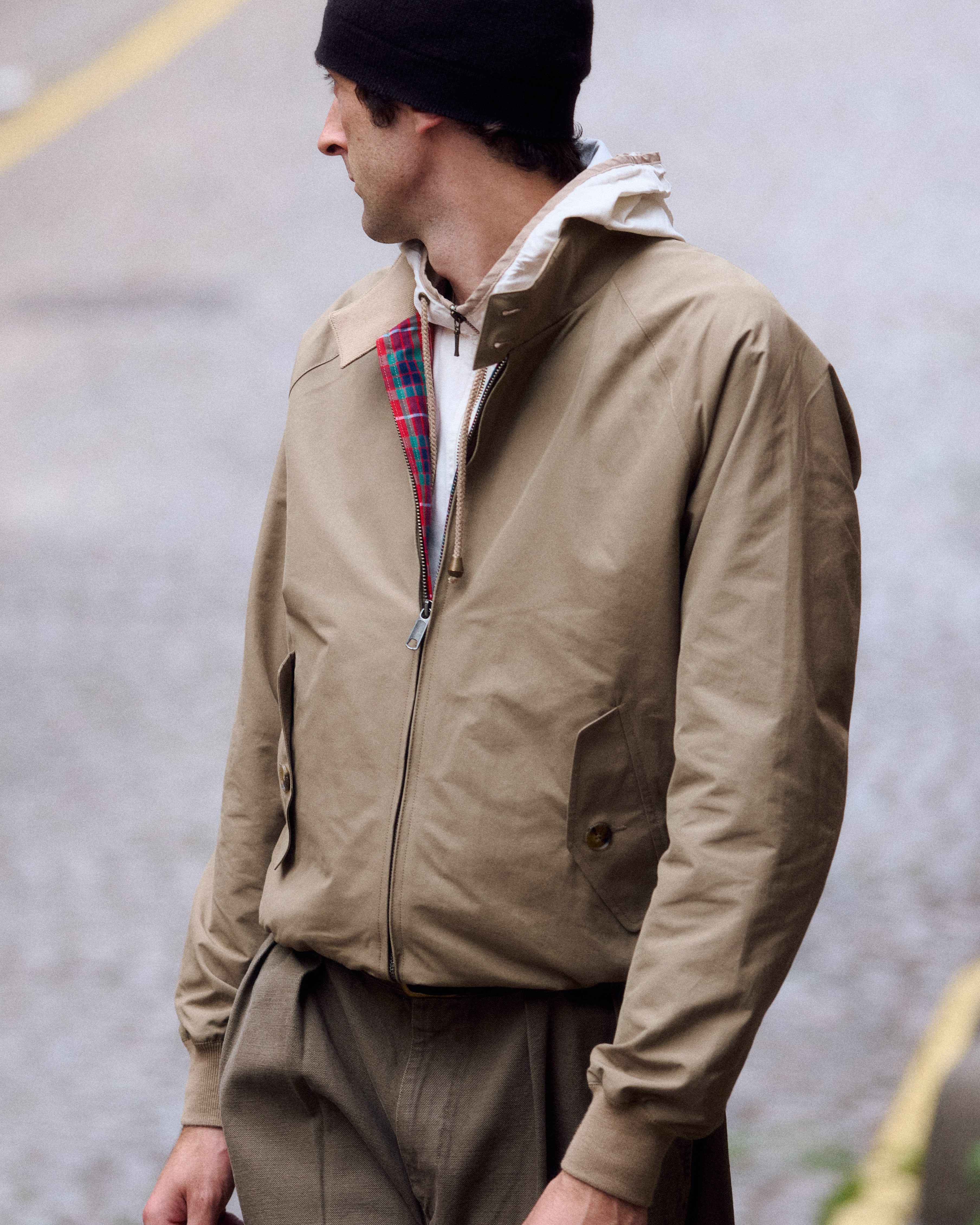 Le mannequin porte un blouson de pluie beige Baracuta et un bonnet noir et tourne le dos à l'objectif