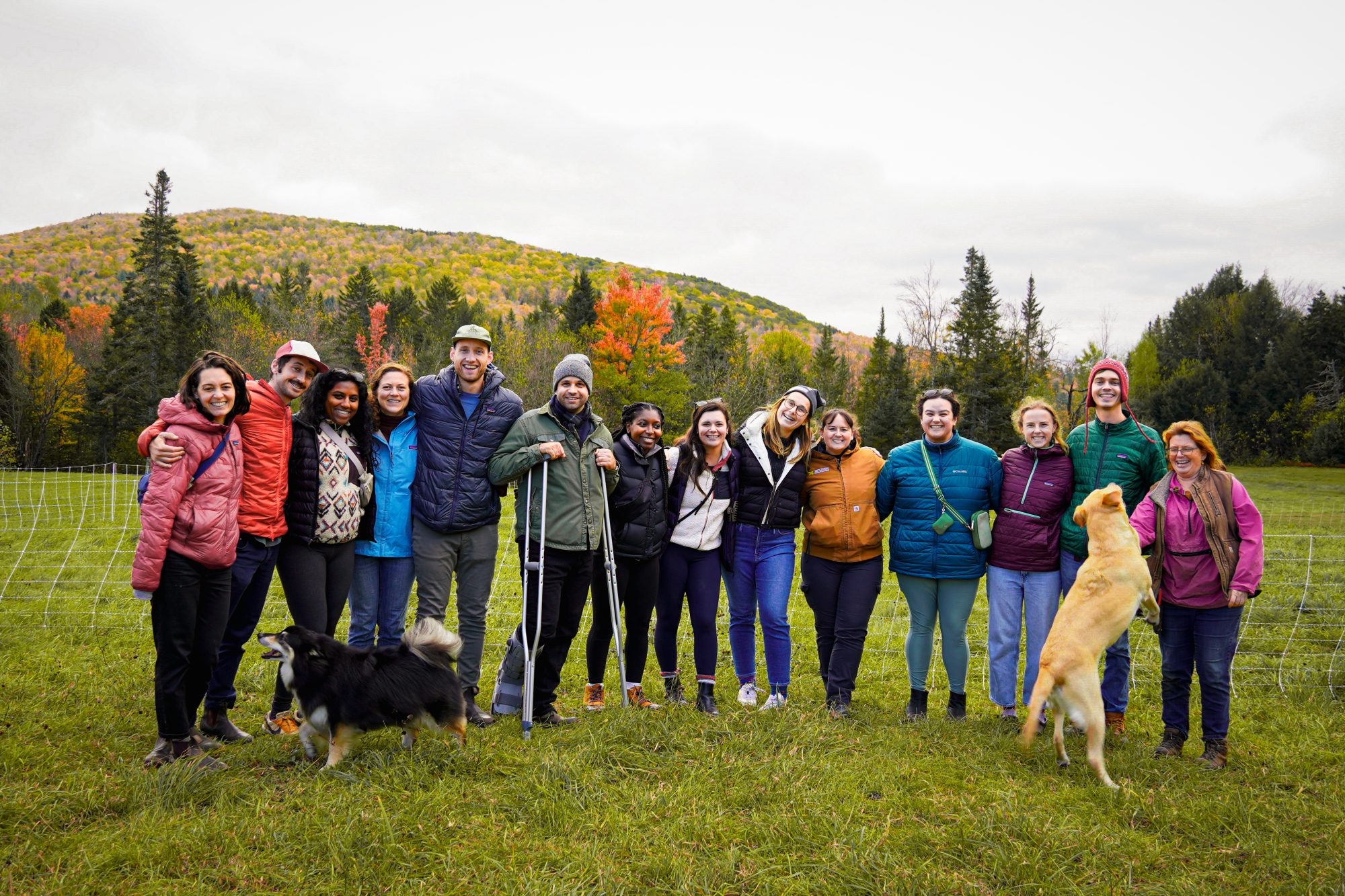 A group of people stands together in a grassy field, with trees and hills in the background, enjoying a day outdoors.