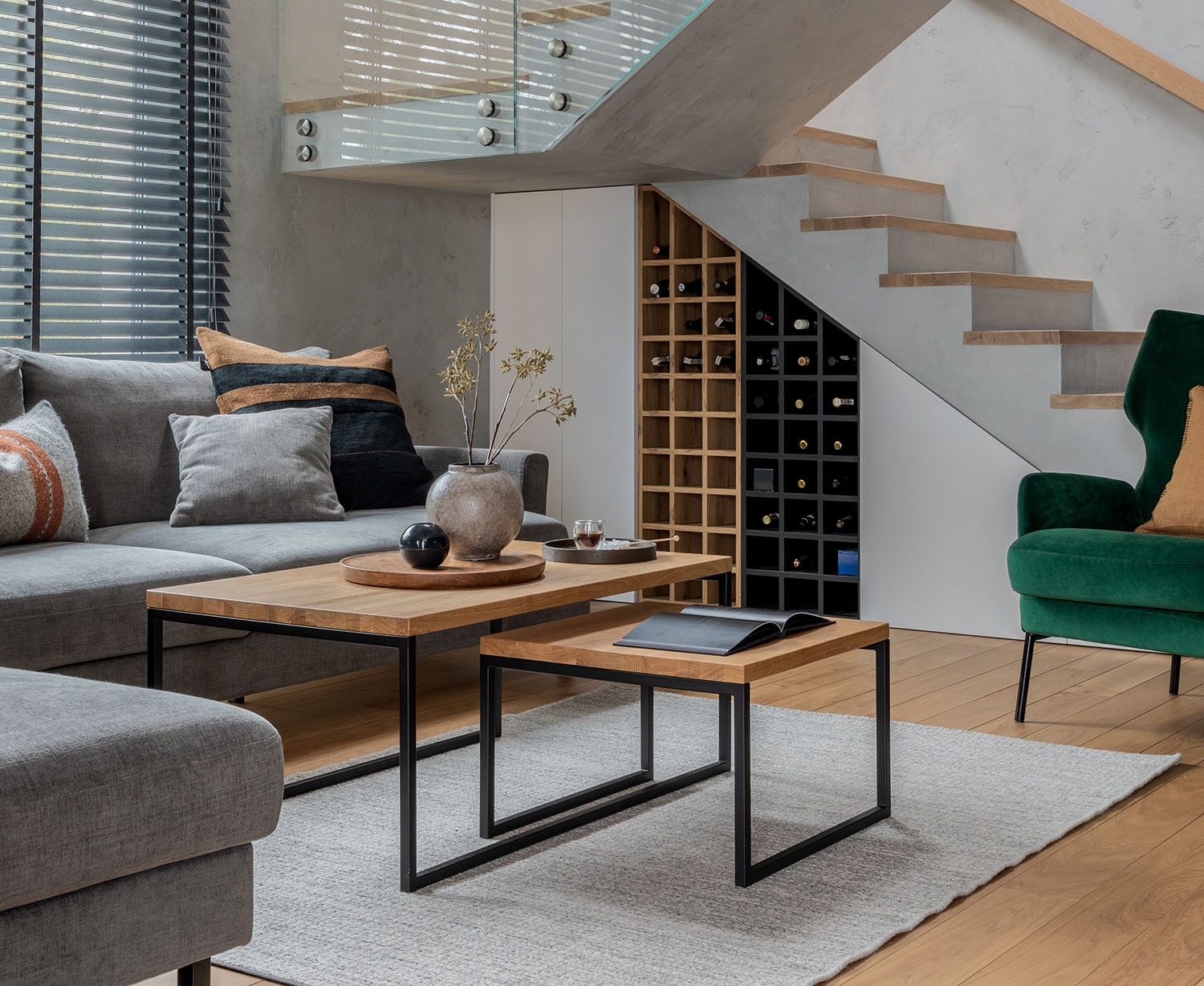 A modern living room with a grey sofa adorned with colorful pillows, a wooden coffee table with decor, a green armchair, and a wine rack under a staircase, set against large windows with blinds.