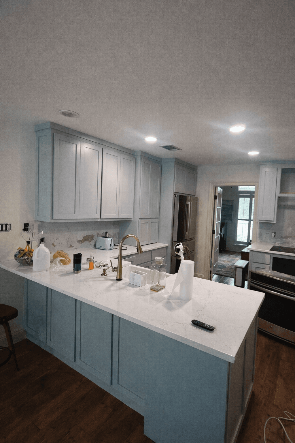 A modern, glass-walled house with warm interior lighting is perched on a rocky cliff overlooking a calm sea at sunset, under a clear blue sky.