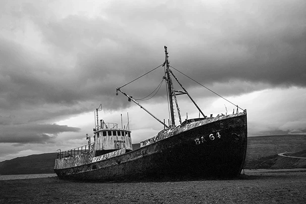 Czarno-białe zdjęcie porzuconego, zardzewiałego kutra rybackiego stojącego na brzegu pod dramatycznym, pochmurnym niebem