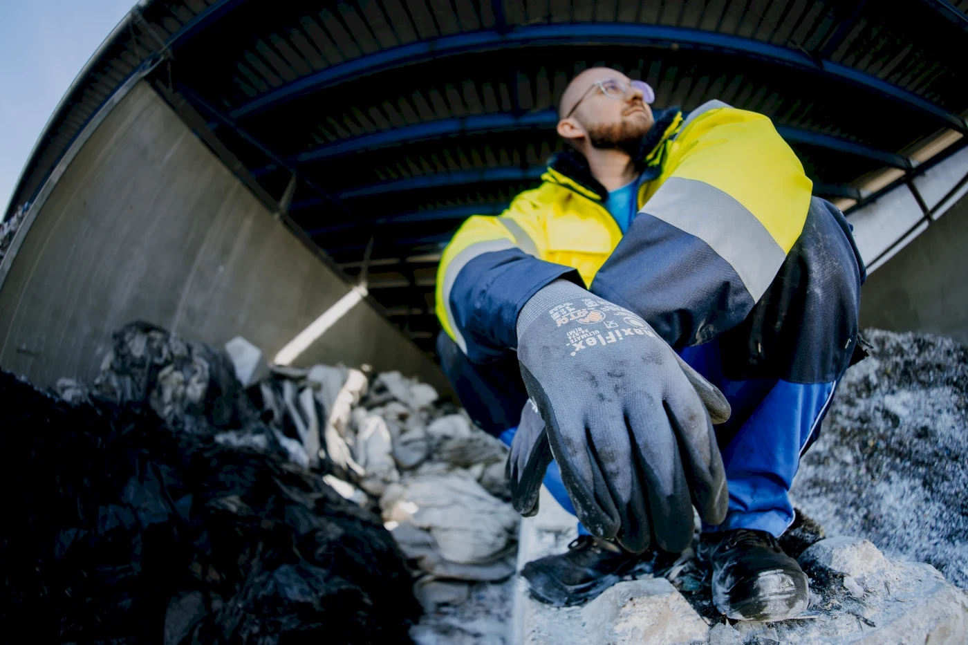 Mann in Sicherheitskleidung sitzt auf Abfallberg und blickt nach oben. Handschuhe, Jacke, Hose, Arbeitsschutz.