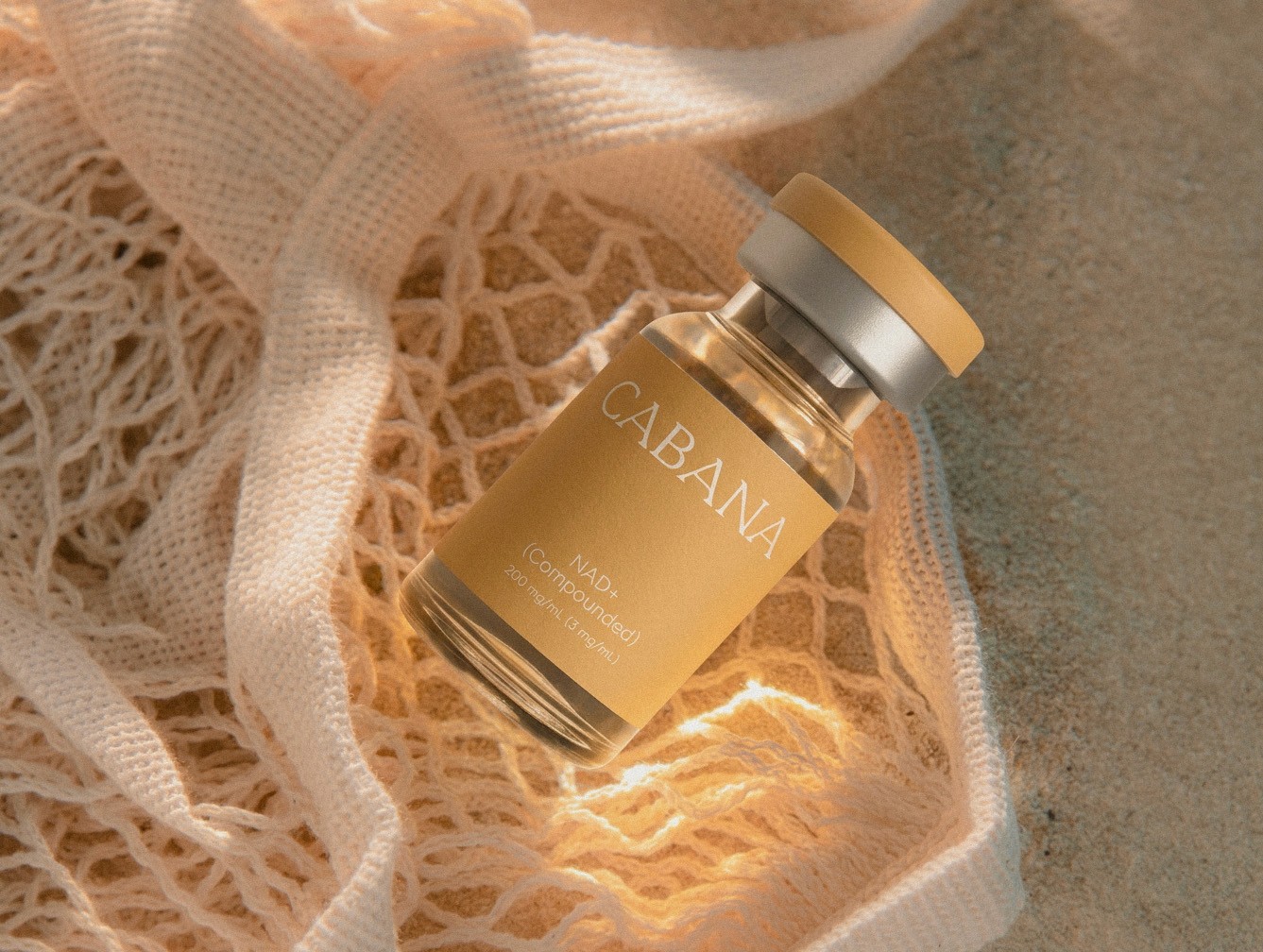 Cabana NAD+ vial resting on a sunlit net bag on sand, warm beach light creating soft shadows and a relaxed, coastal feel