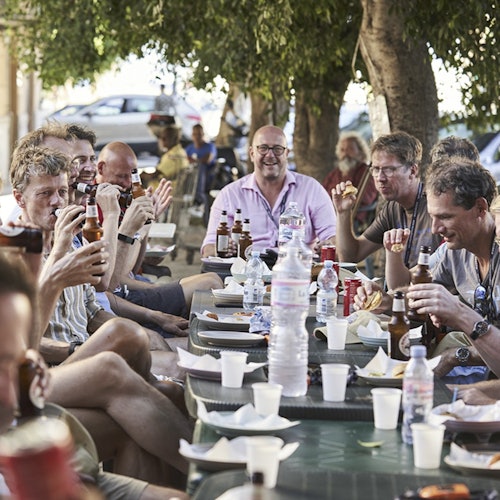 一群男人坐在戶外長桌旁，喝著啤酒，吃著東西，在大樹的陰影下笑著。