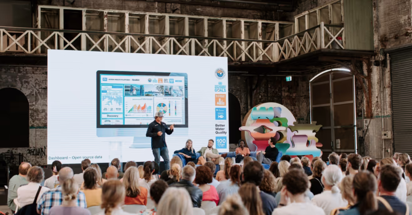 A speaker presenting a data dashboard on a large screen to a seated audience in a warehouse venue, with a colourful sculptural installation beside the stage