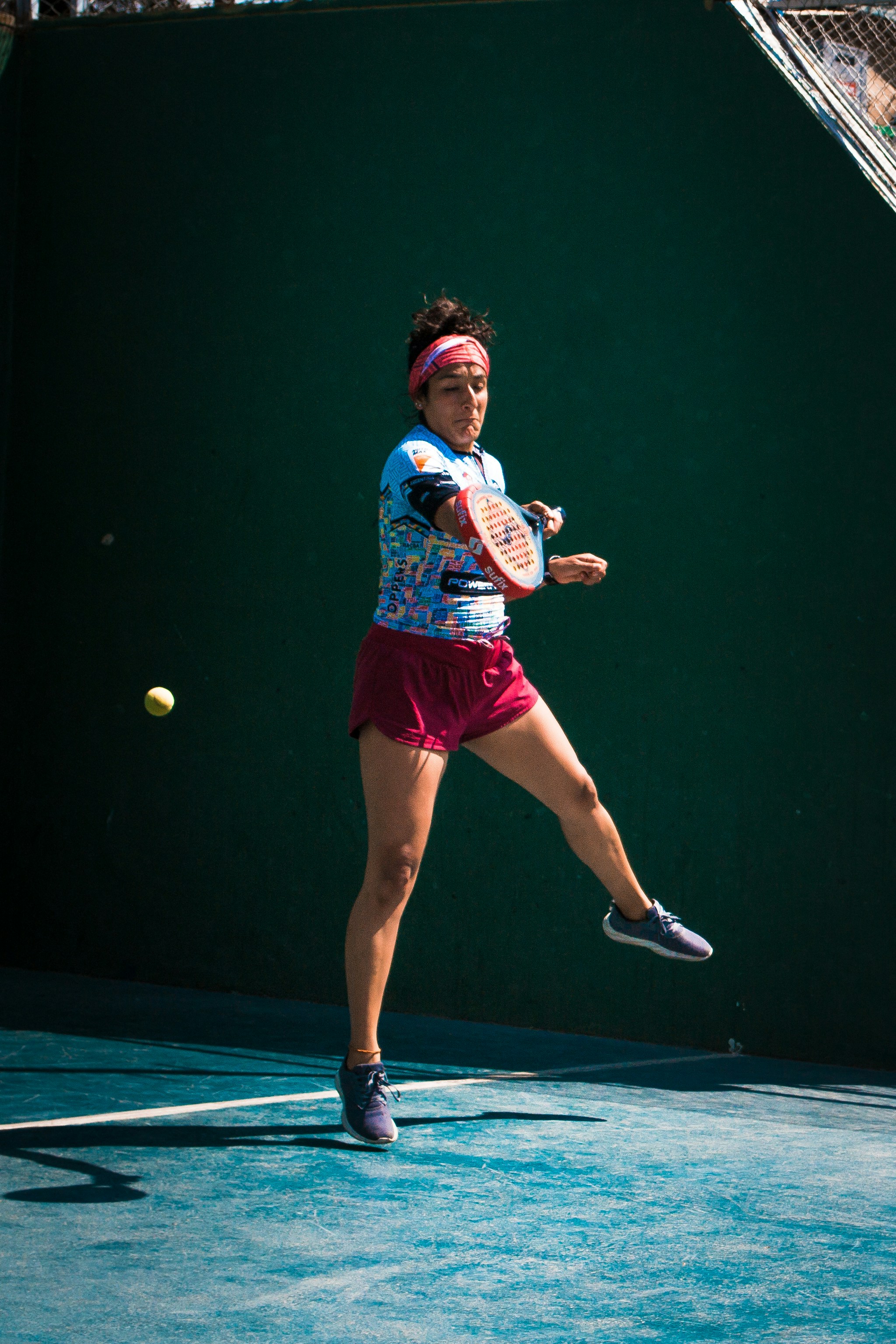 A person in athletic apparel is captured mid-action playing padel, swinging a racket to hit a ball, against a dark green court wall background.