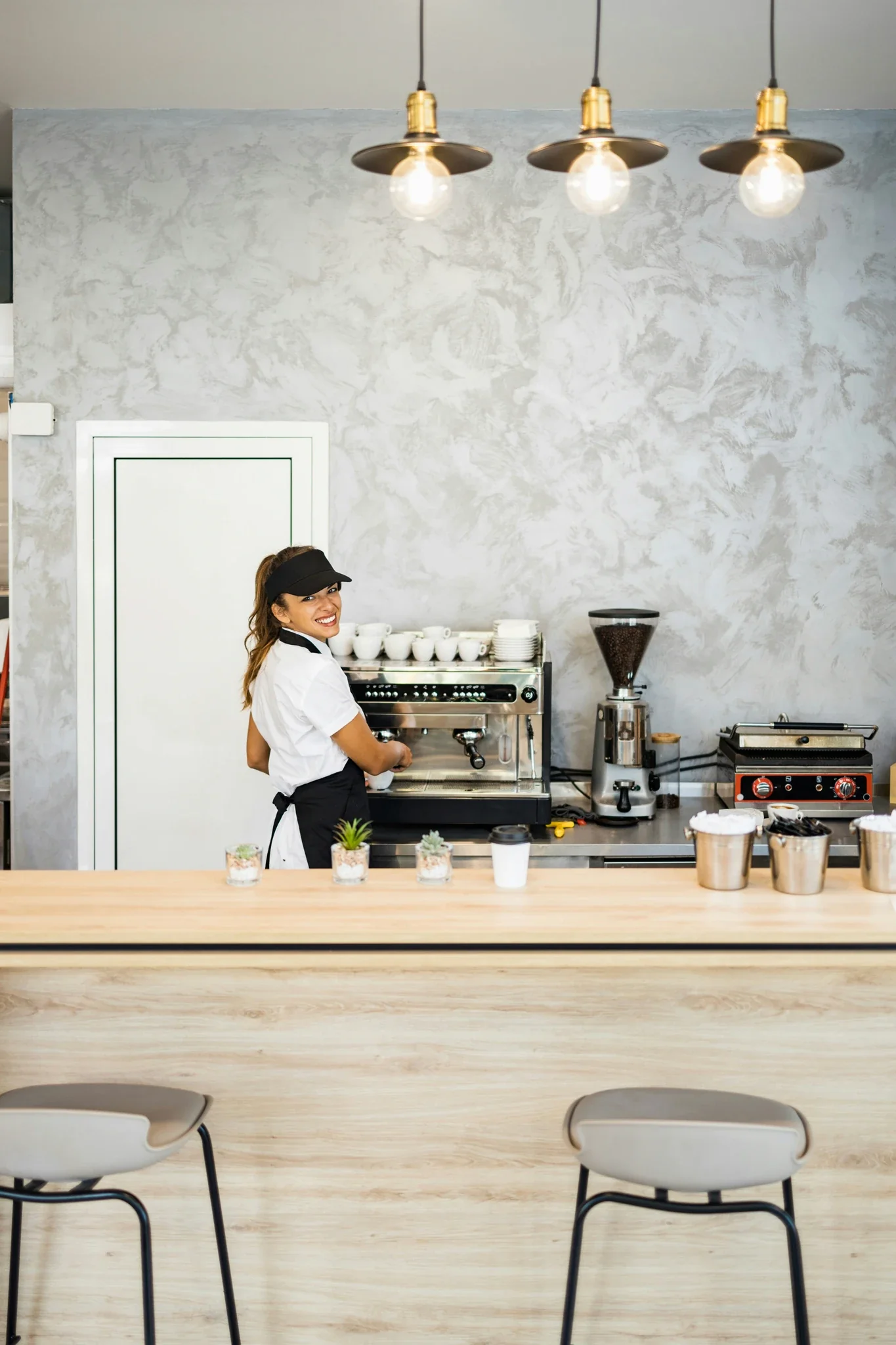 Smiling barista in black cap and white shirt working at espresso machine behind wooden counter in modern café.