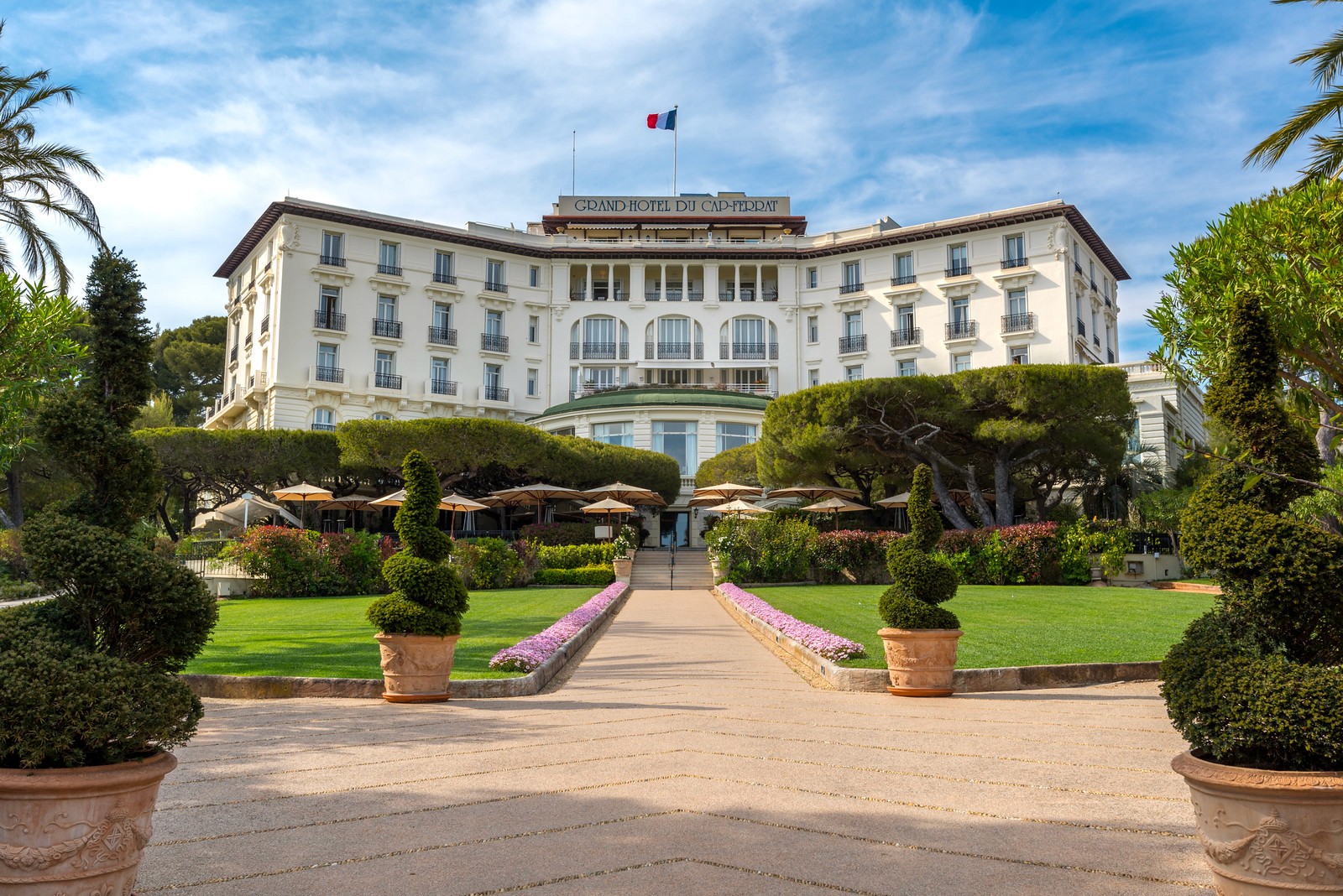 A grand, symmetrical white palace with a French flag flying on top, fronted by manicured gardens and topiaries.