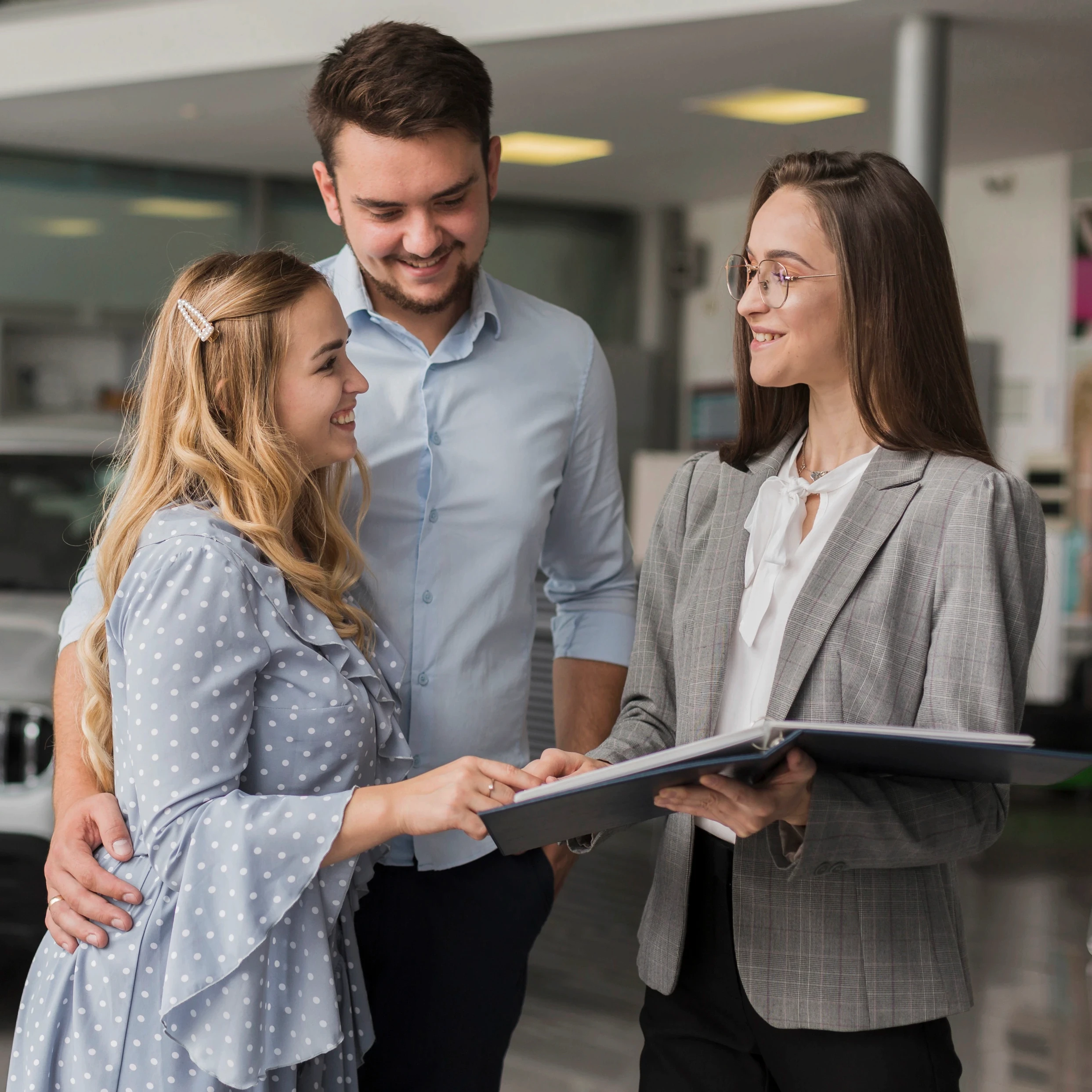 A smiling couple consults with a professional woman holding a folder in an office setting. The mood is positive and collaborative.