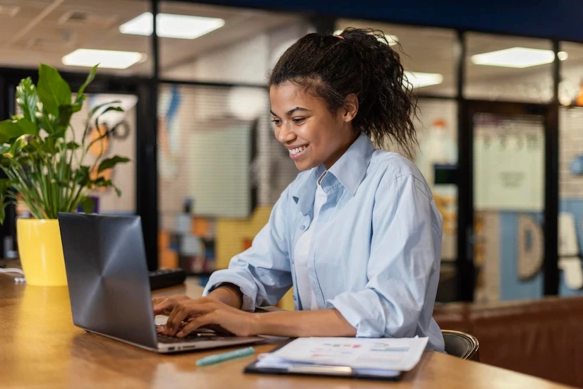 A imagem mostra uma mulher jovem sorrindo enquanto trabalha em seu laptop. Ela está sentada em uma mesa, com papéis e uma caneta ao lado, sugerindo que está envolvida em uma atividade profissional ou acadêmica. A mulher veste uma camisa azul clara e parece estar em um ambiente de escritório moderno, com divisórias de vidro ao fundo e plantas decorativas na mesa. A cena transmite uma atmosfera positiva e produtiva, destacando uma mulher concentrada em seu trabalho em um ambiente acolhedor e organizado.