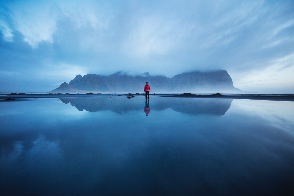 A man looking at a mountain in the distance standing on a wet stretch of land on a cloudy day