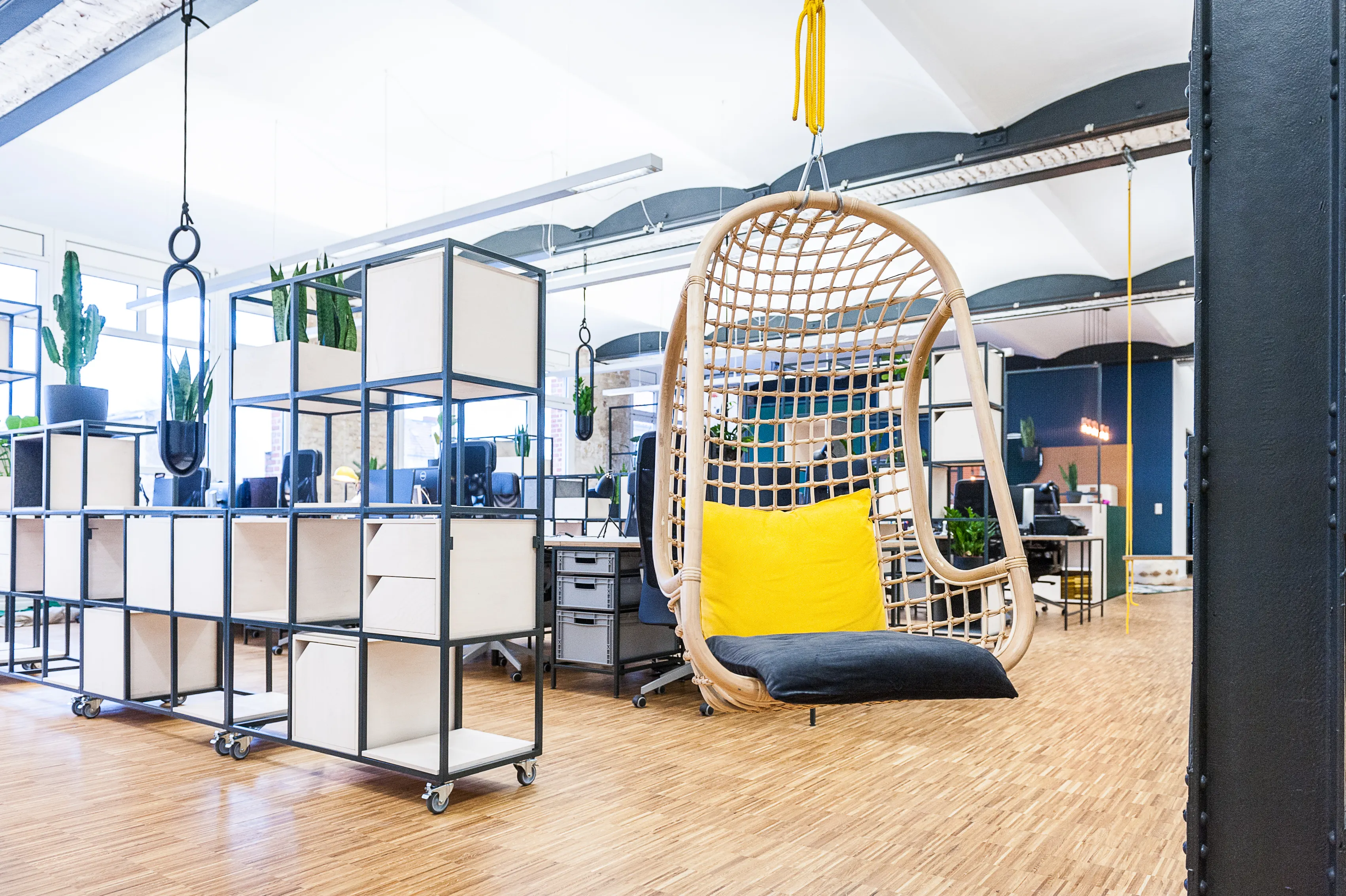 Hanging rattan egg chair with yellow and dark blue cushions in the open-plan office of INTERMATE x TRUEMATES, surrounded by white modular shelves, cacti and desks on oak parquet.