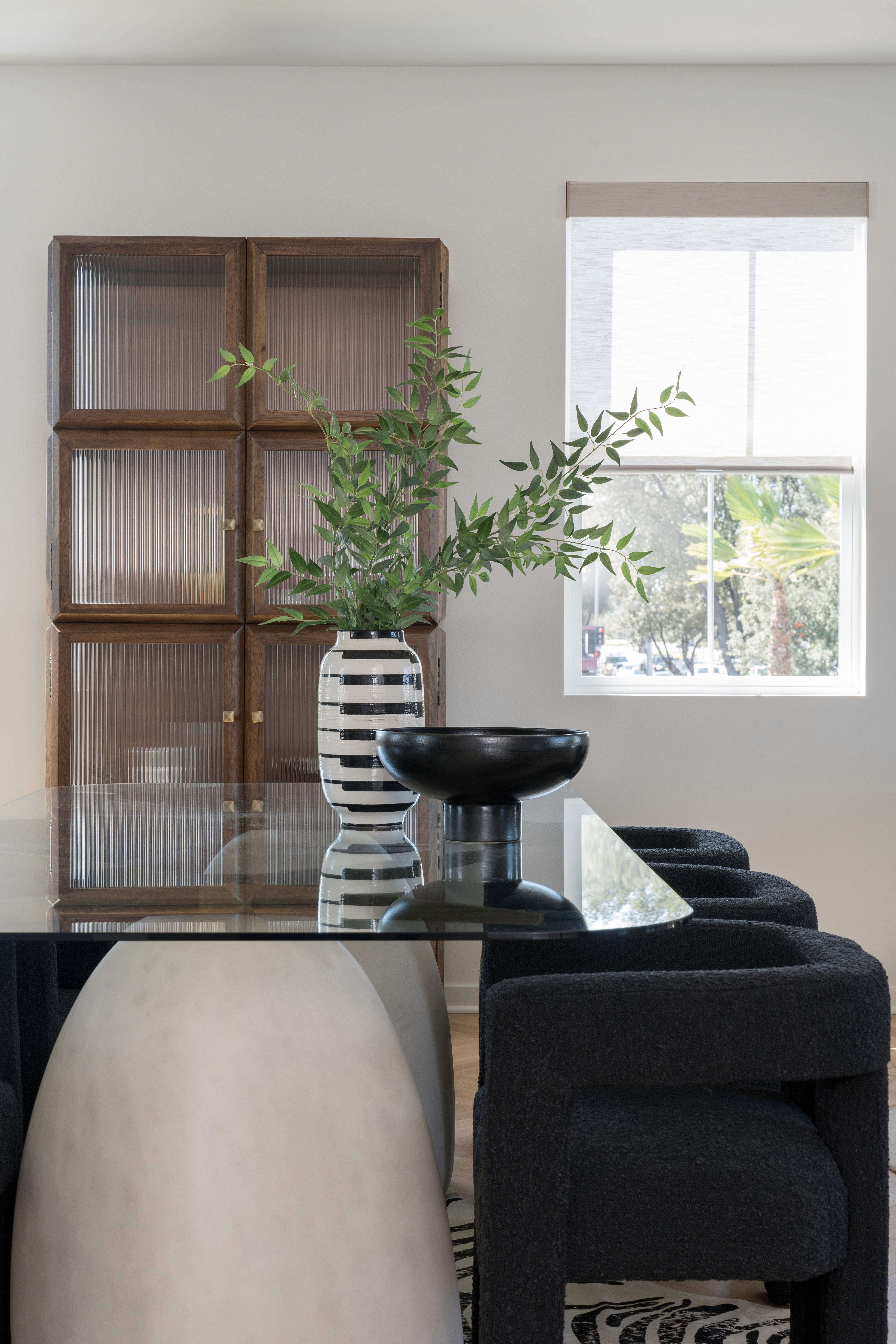 Dining area with black boucle dining room chairs, modern, glass top dining table, black pedestal bowl and black and white striped vase; wood storage cabinet with glass doors in background