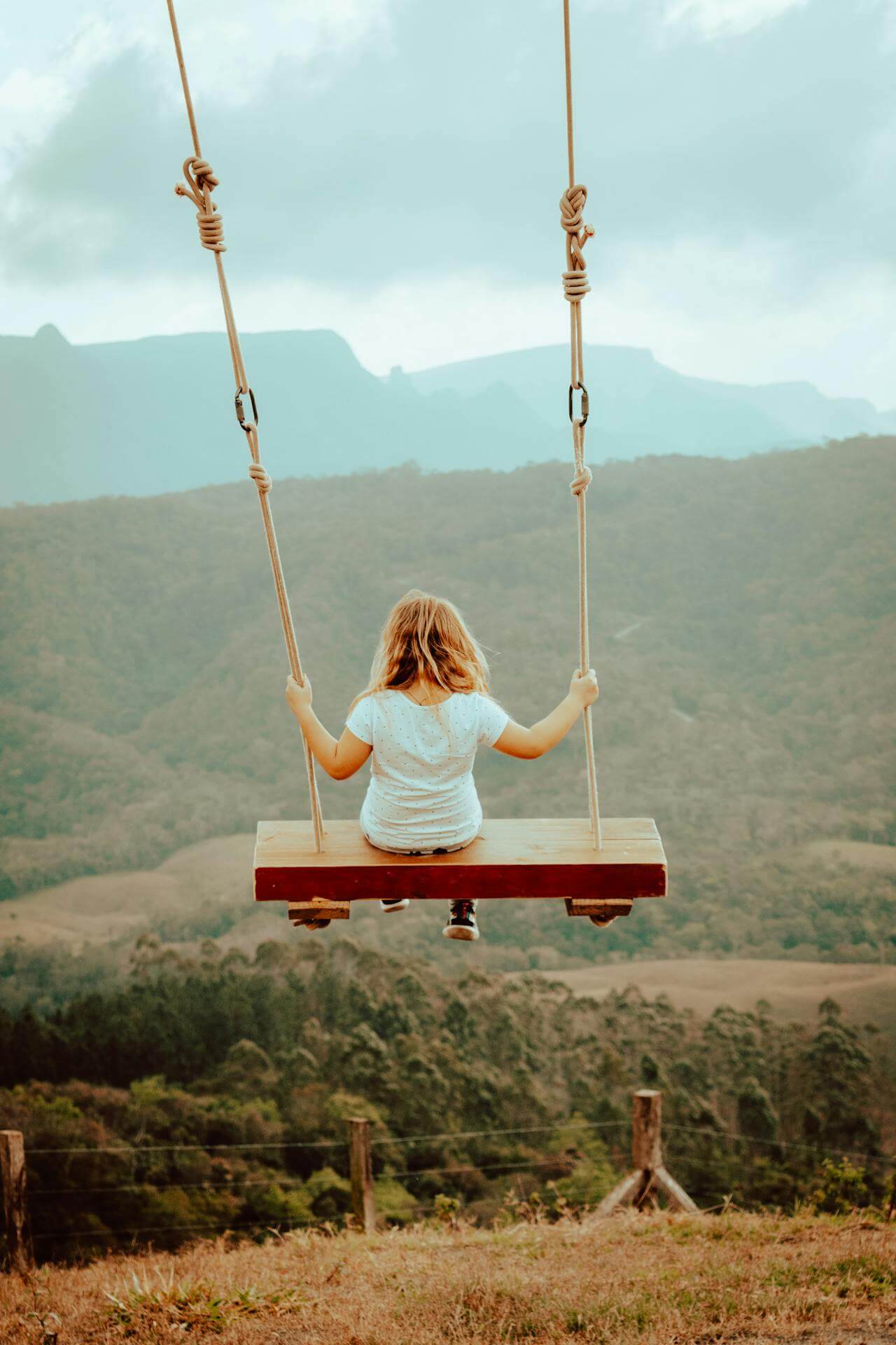 A young girl is seated on a swing, overlooking a vast, serene landscape of mountains and forests.