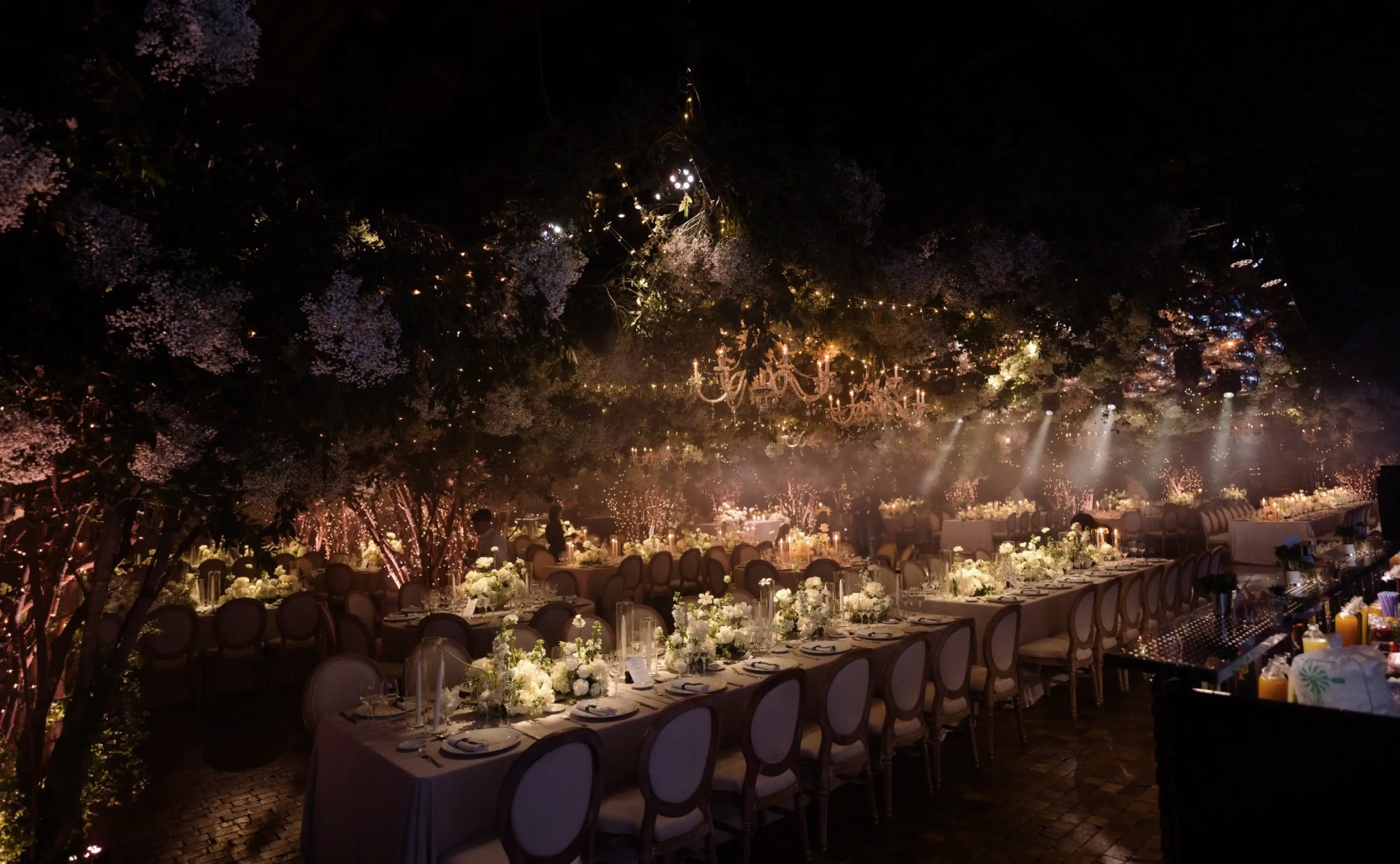 Salón iluminado con candelabros, mesas largas y una ambientación floral blanca bajo un techo de follaje.