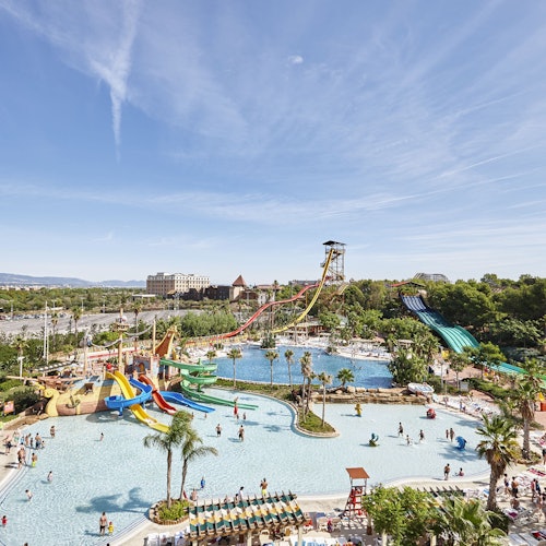 A vibrant water park with multiple slides, pools, and palm trees under a clear blue sky; people are enjoying the attractions.