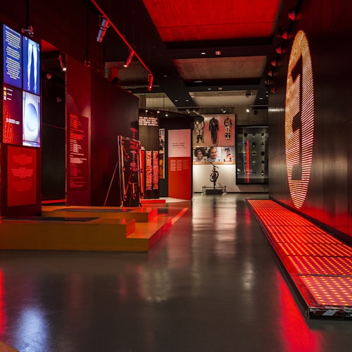 Mostra museale moderna con illuminazione rossa, esposizioni di attrezzature sportive, pannelli informativi e un grande numero 3 illuminato.