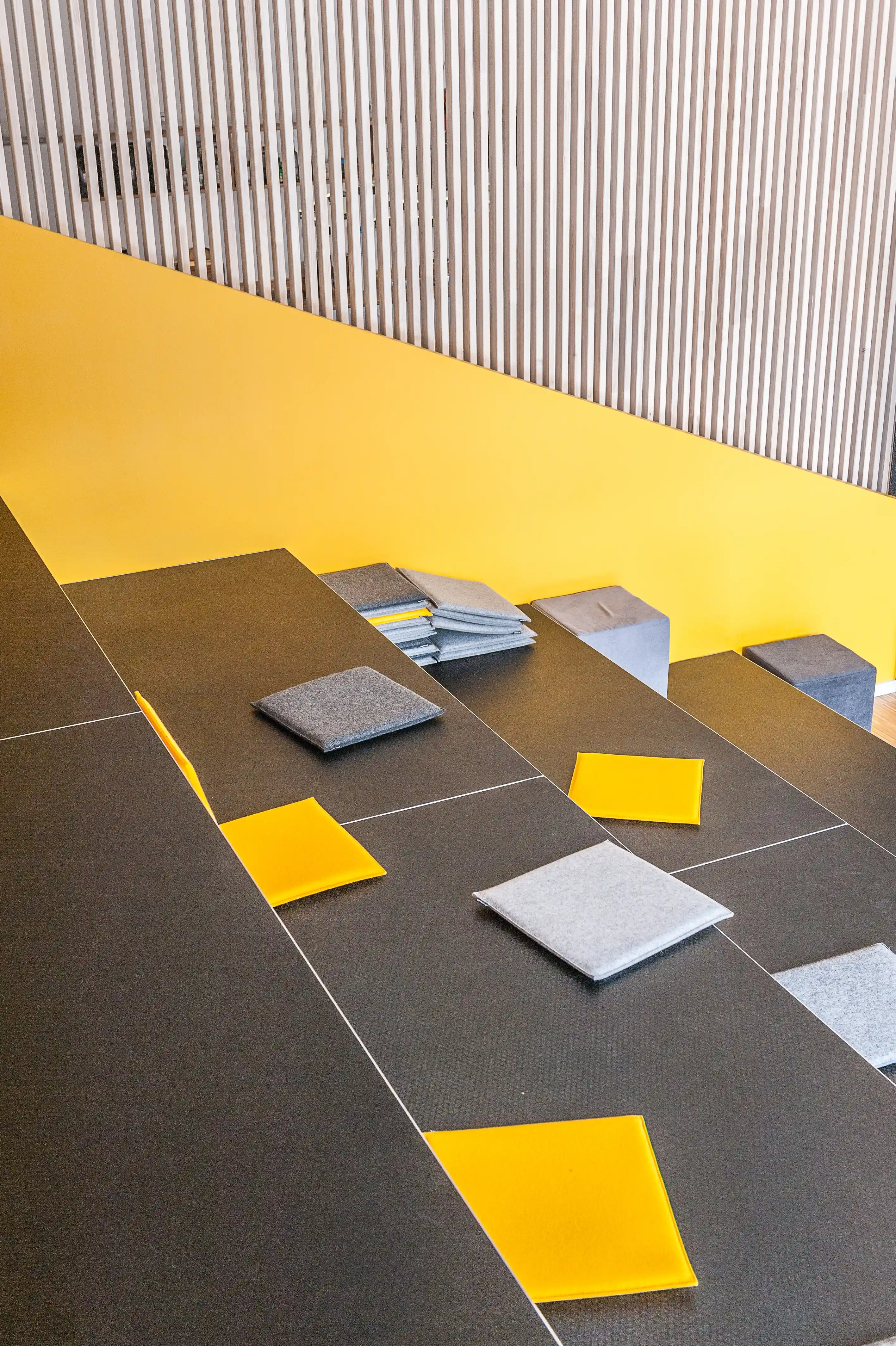Detail of the dark grey seating tribune at BASILICOM with yellow and grey felt cushions, folded blankets and concrete-look stools against a bright yellow wall.
