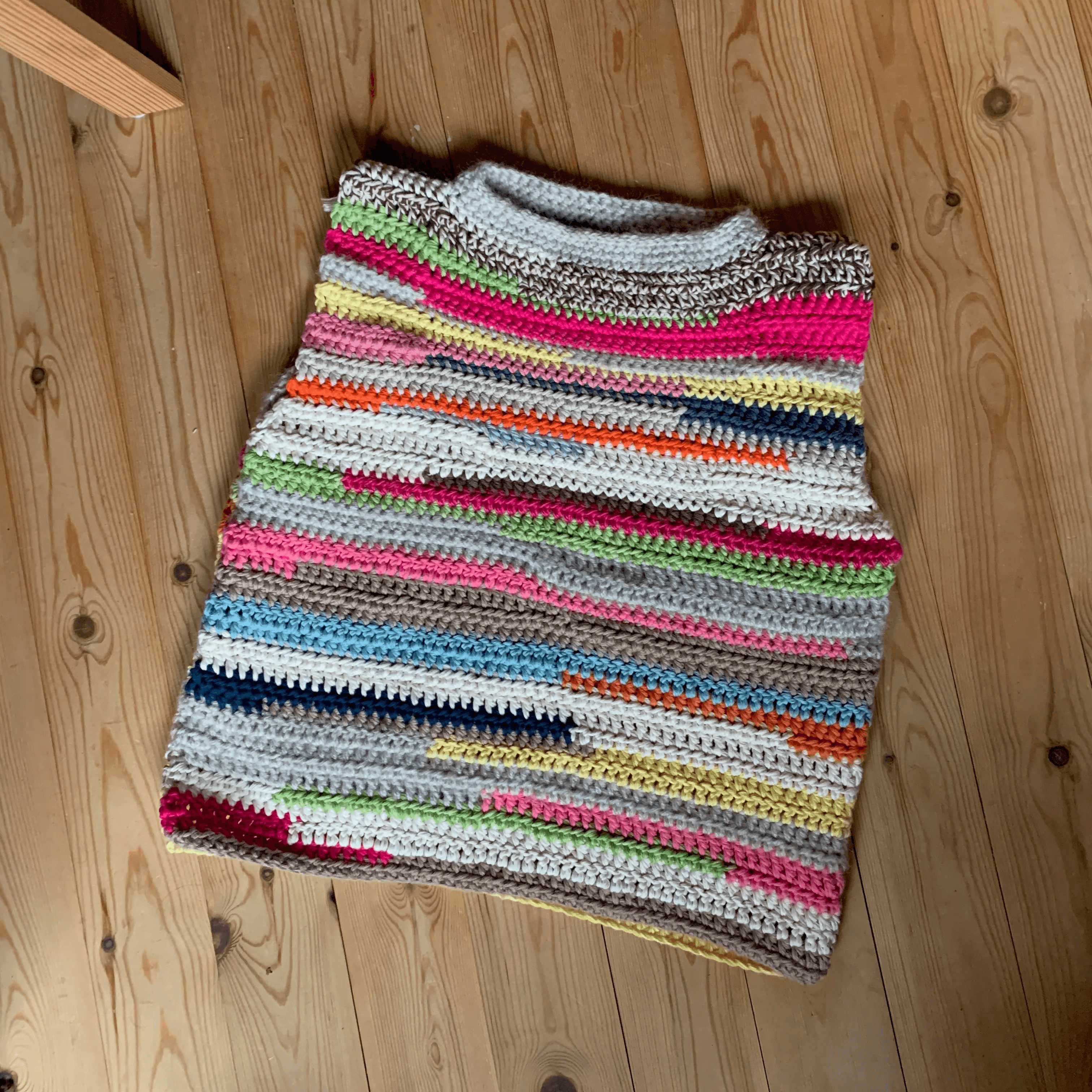 A colorful, handmade crochet vest featuring a variety of horizontal stripes in vibrant hues lies neatly on a wooden floor, showcasing intricate texture and craftsmanship.