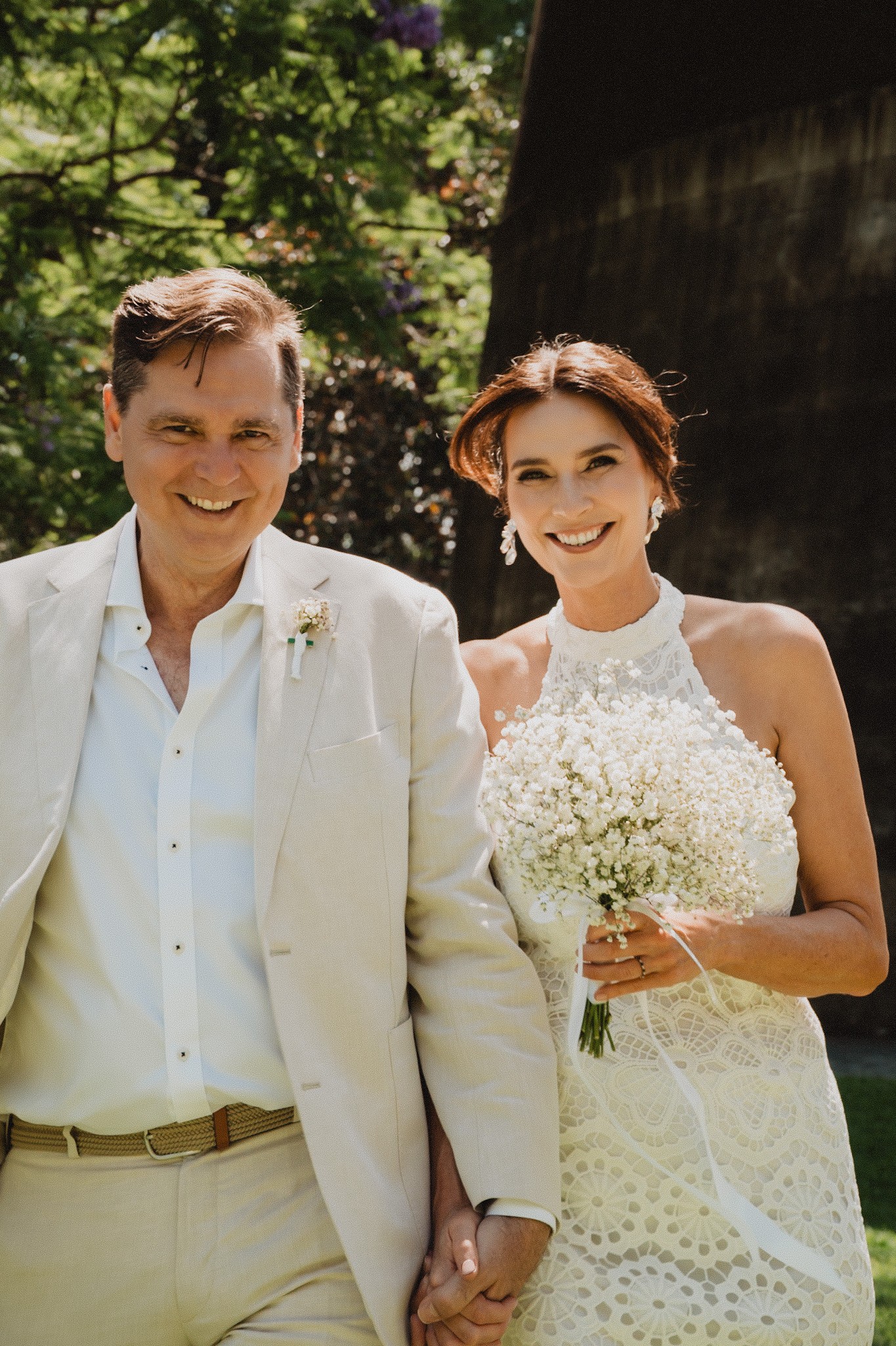 Smiling bride and groom holding hands, with a white flower bouquet and sunlit greenery behind them.