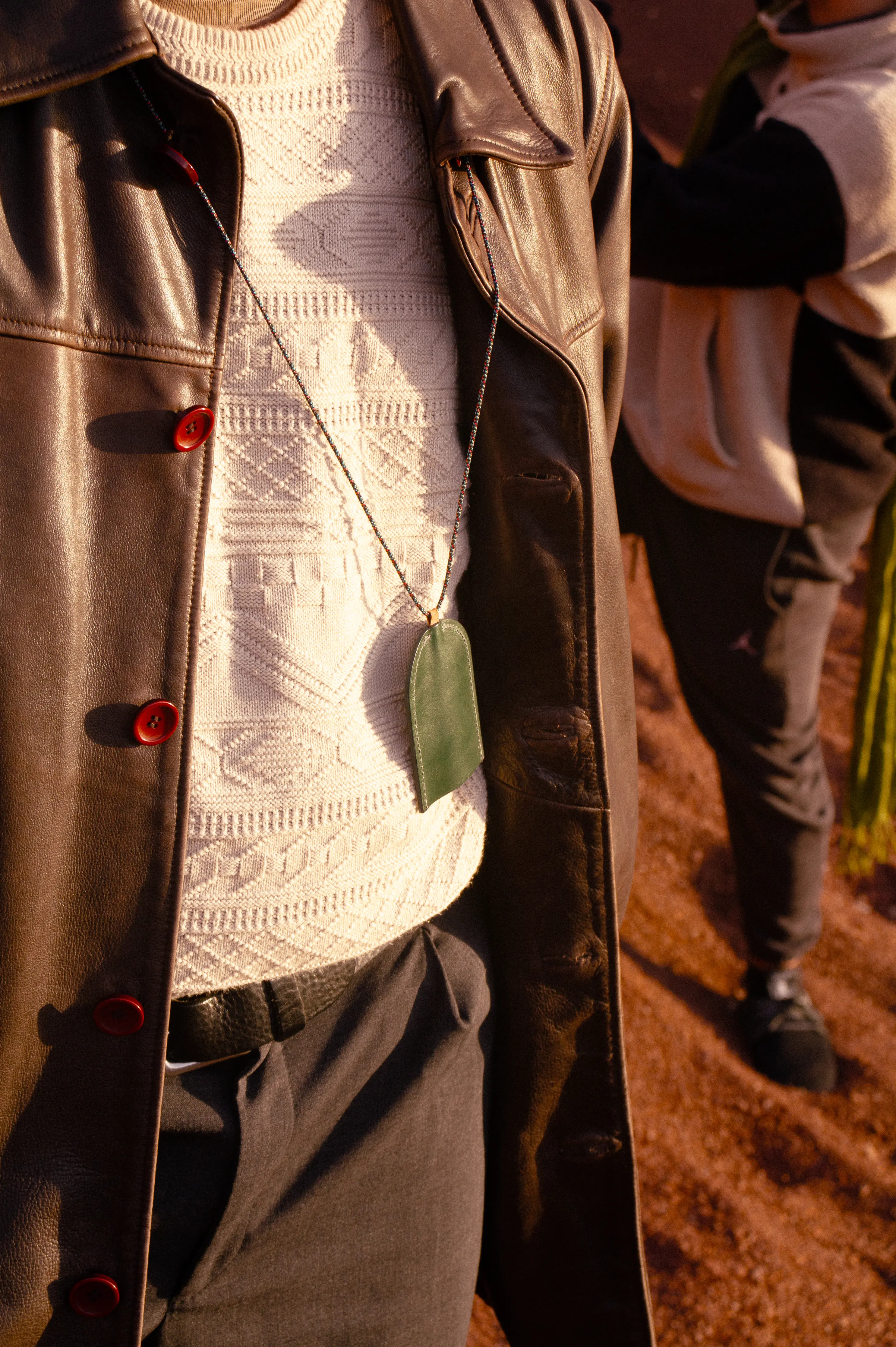 Close-up of a brown leather coat over a textured off-white sweater, with a green leather pendant, while a second figure appears blurred in the background.
