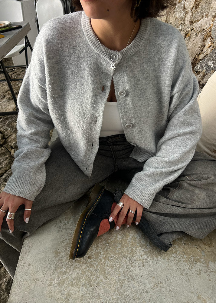 Person wearing a gray cardigan sitting on a stone surface. Wearing a silver necklace.