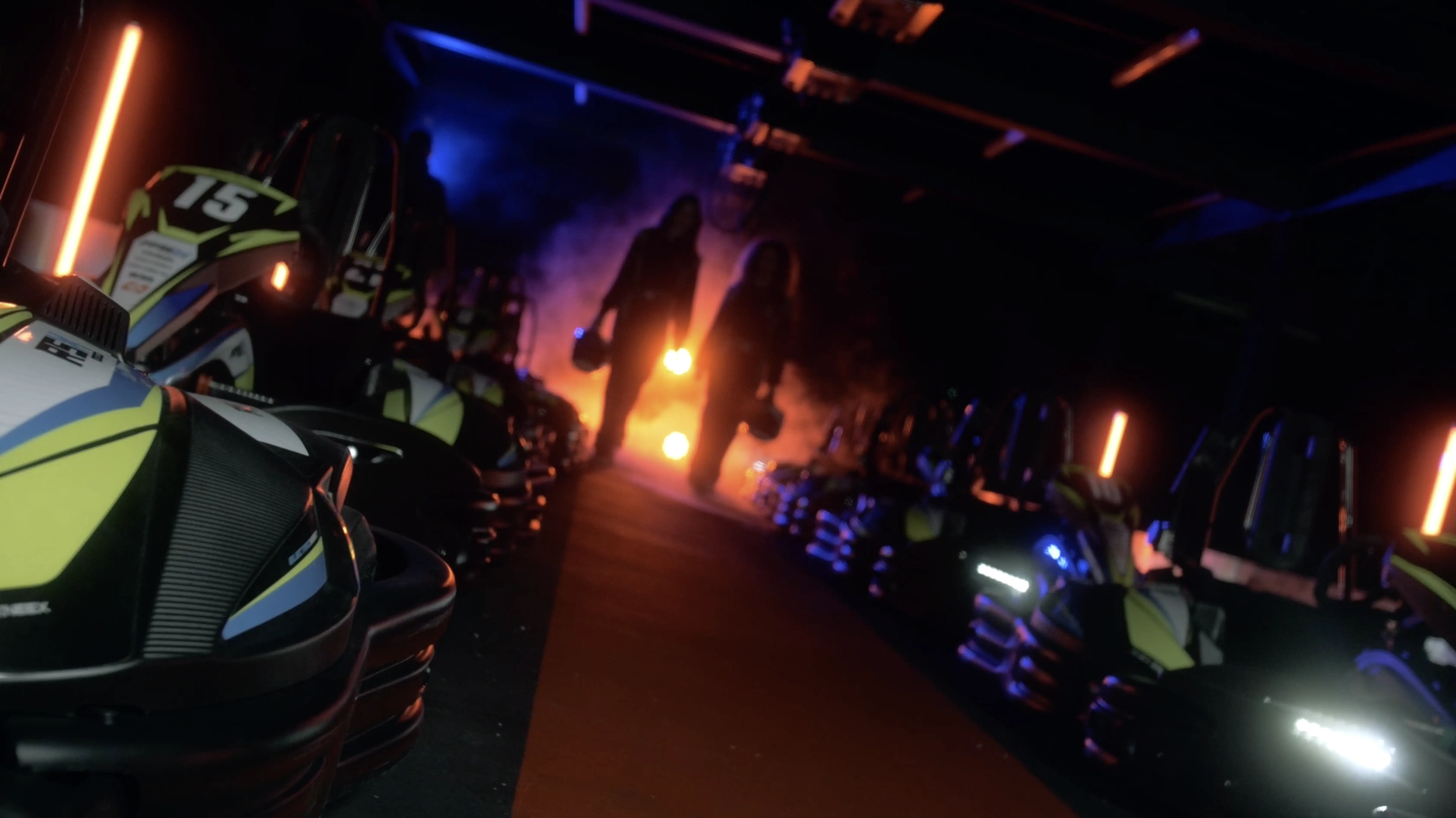Two people carrying helmets walk down a dimly lit go-kart track, surrounded by neatly lined go-karts and illuminated by vibrant blue and orange lights.