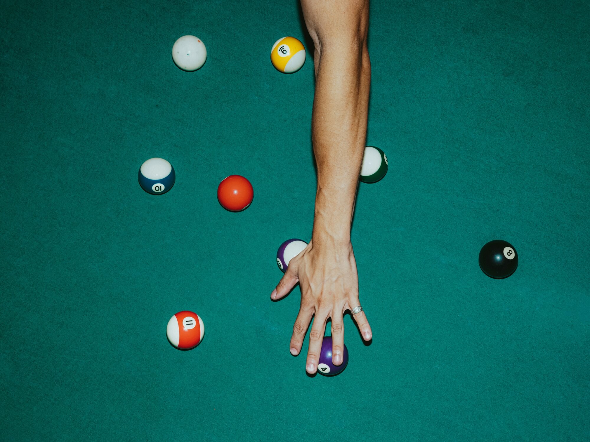 A person's hand on a pool table, fingers spread over a purple billiard ball. Several other colorful balls are scattered on the green felt surface.