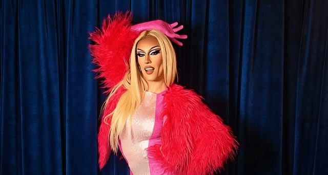 A drag performer with vibrant blue wavy hair and dramatic makeup looking at her reflection in a large mirror, wearing a distressed denim two-piece outfit.