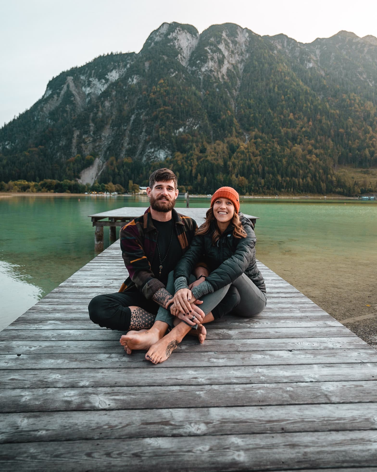 Bre and Flo sit together on a wooden dock by a tranquil lake, surrounded by mountains. The woman wears an orange beanie, both are smiling.