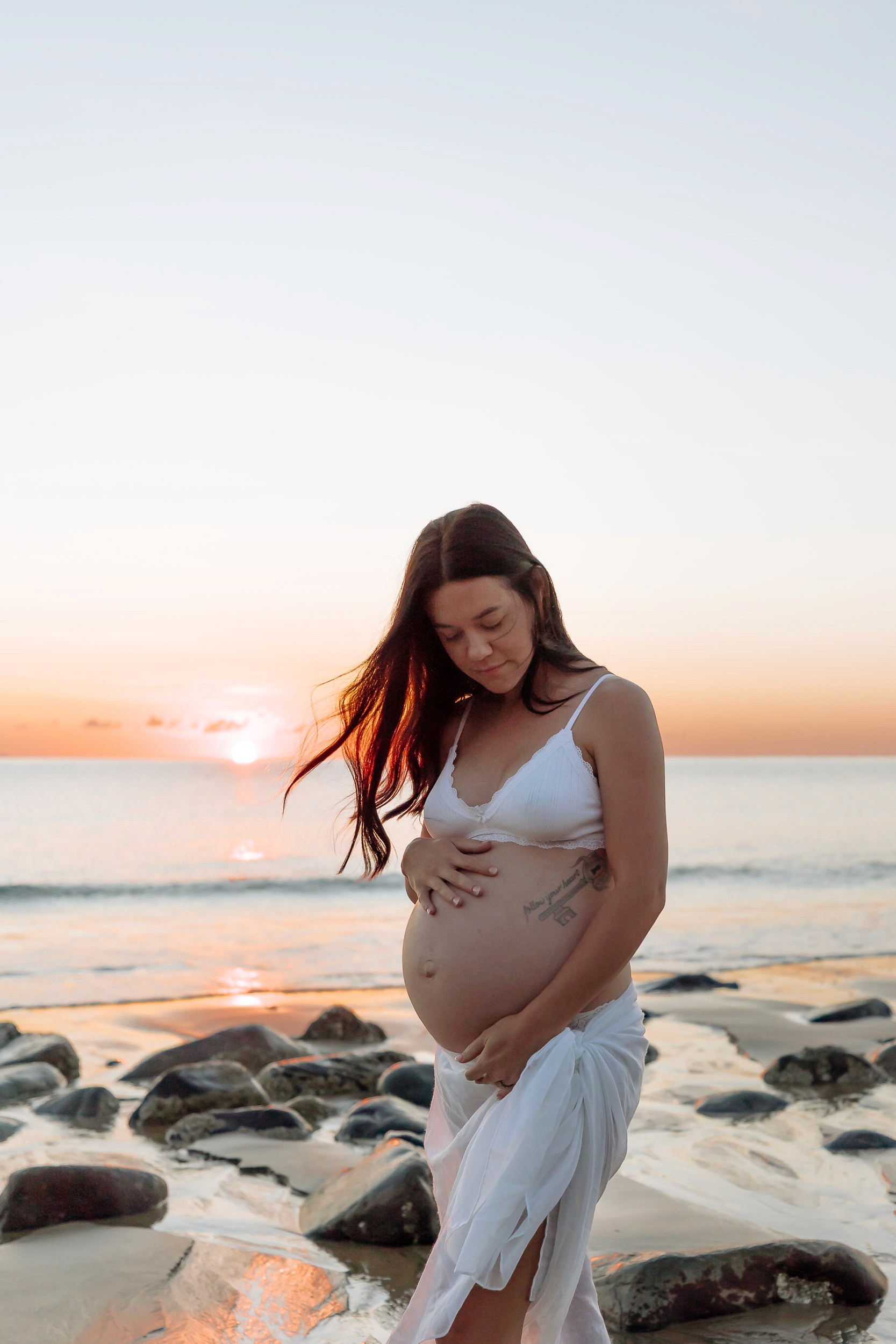 Solo maternity session at sunrise on the beach, pregnant woman in a flowing skirt standing barefoot at the water’s edge as soft golden light reflects on the ocean in Mackay.