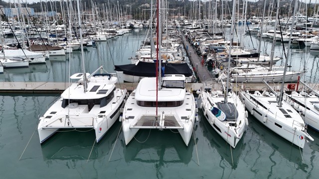 Elevated aerial view of a Lagoon 42 sailing catamaran with solar panels on the coachroof berthed between two other catamarans at a large Mediterranean marina, with rows of sailing yachts and a busy pontoon walkway stretching into the background under an overcast sky