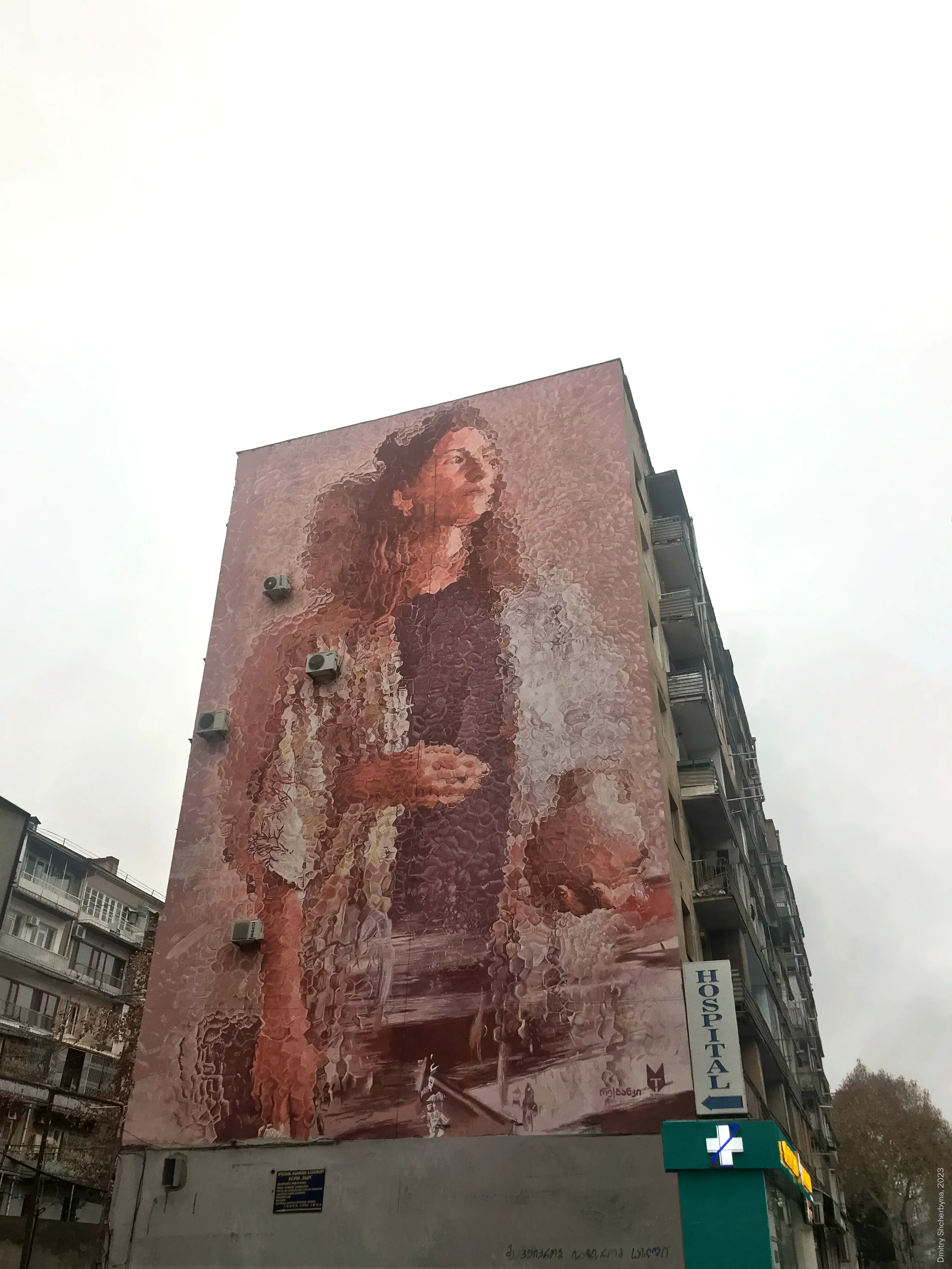 A full-building mural of a woman with long, curly hair rendered in a textured monochromatic palette of pink and mauve, looking upward with her hands gently clasped.
