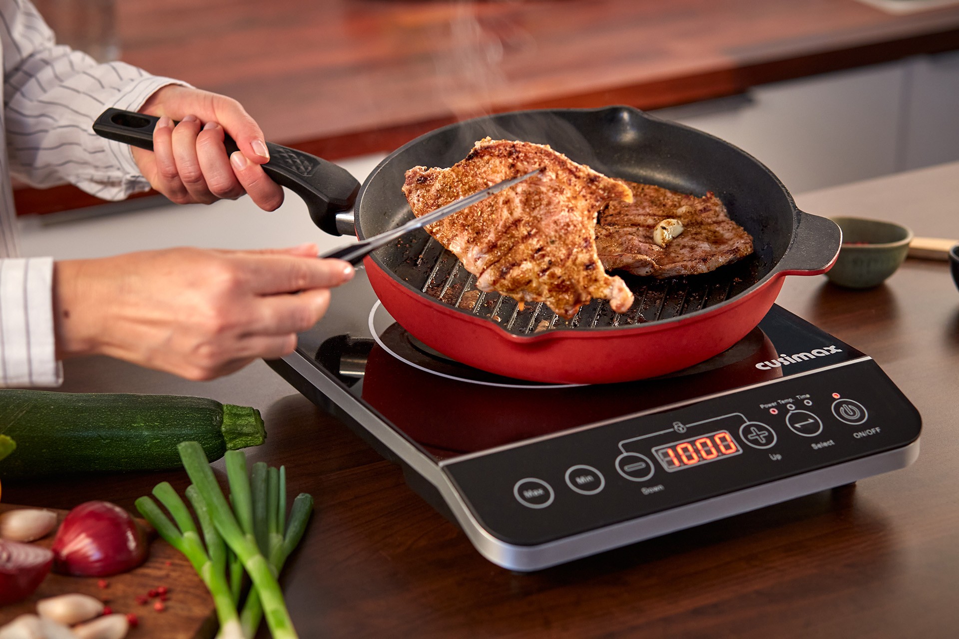 A person grills seasoned meat in a red grill pan on an induction cooktop, with fresh vegetables like zucchini, green onions, and garlic nearby on a wooden kitchen counter.