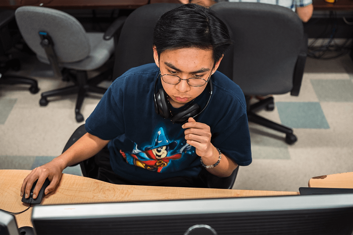 A young person, wearing glasses and a graphic t-shirt, sits focused in front of dual computer monitors while using a mouse, in a room filled with office chairs and desks.