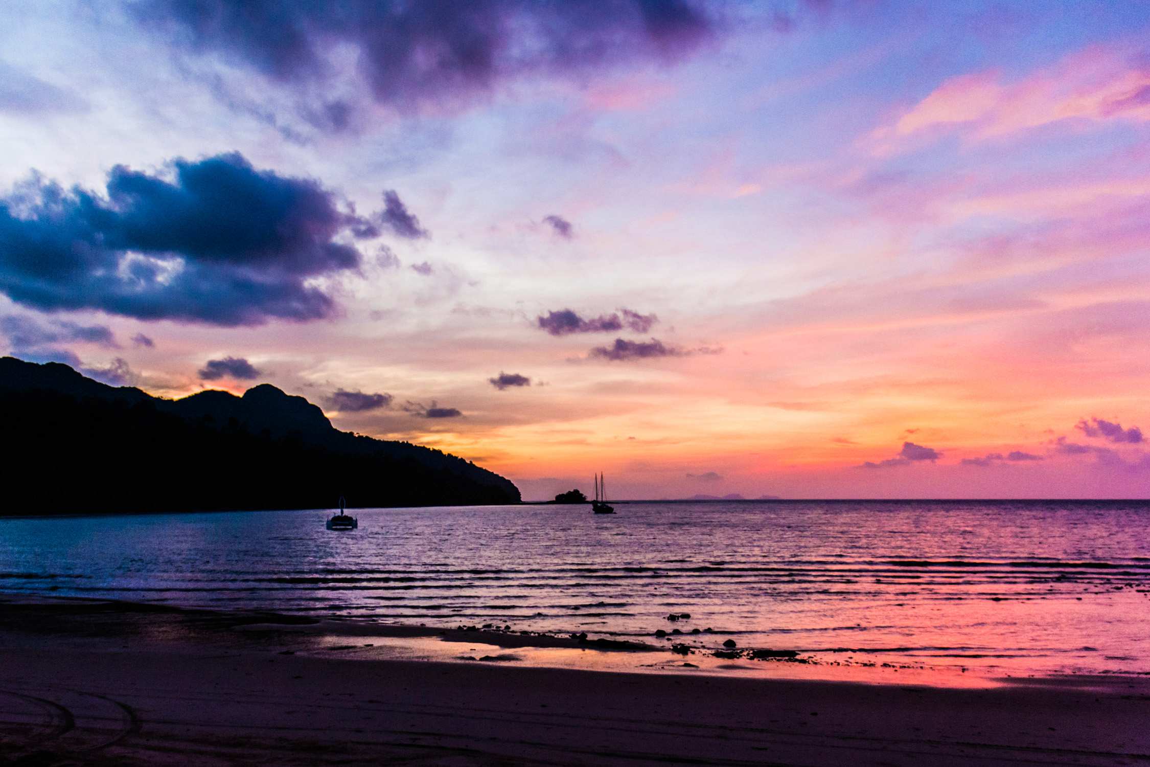A beach at sunset with pink and orange clouds over a calm sea, silhouettes of boats, and dark rocky hills.