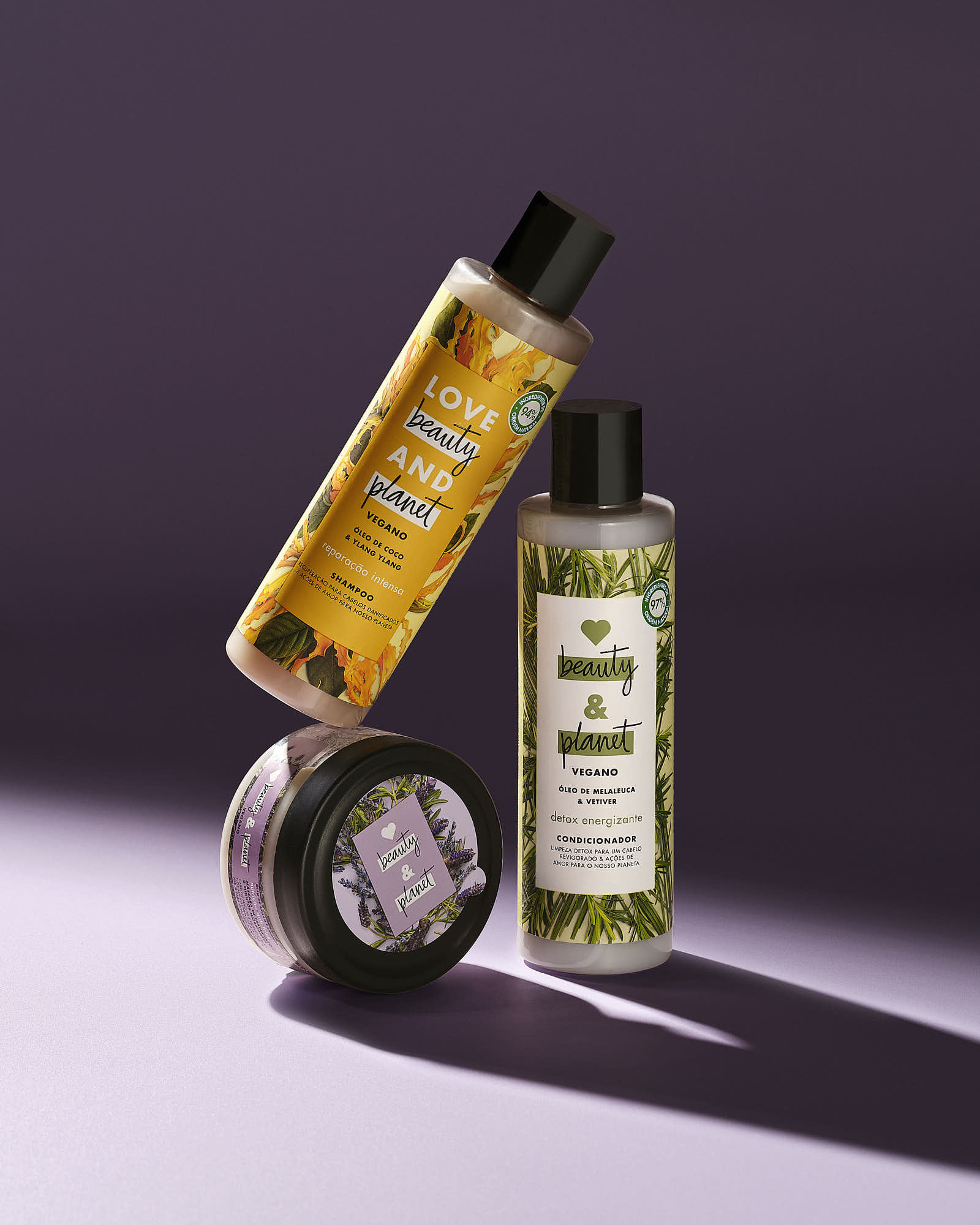 Two bottles of shampoo stand upright next to a round container, casting shadows on a soft, purple background.